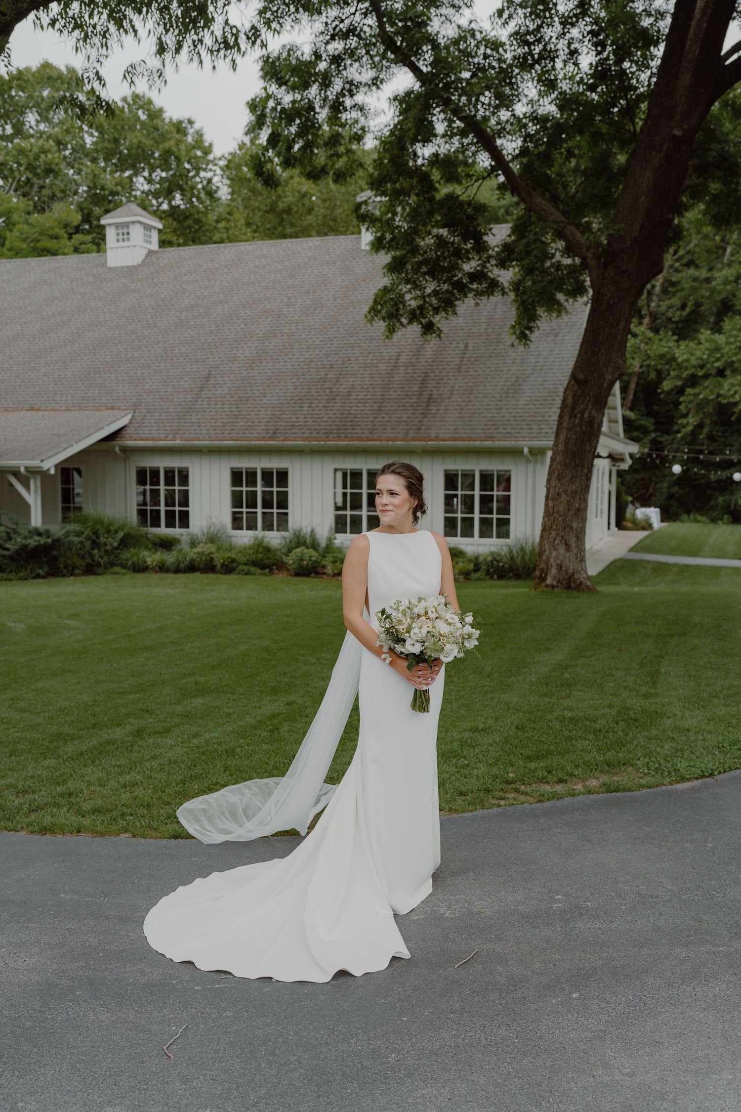 A bride walks gracefully on a curved driveway, her white dress flowing behind her near a classic building.