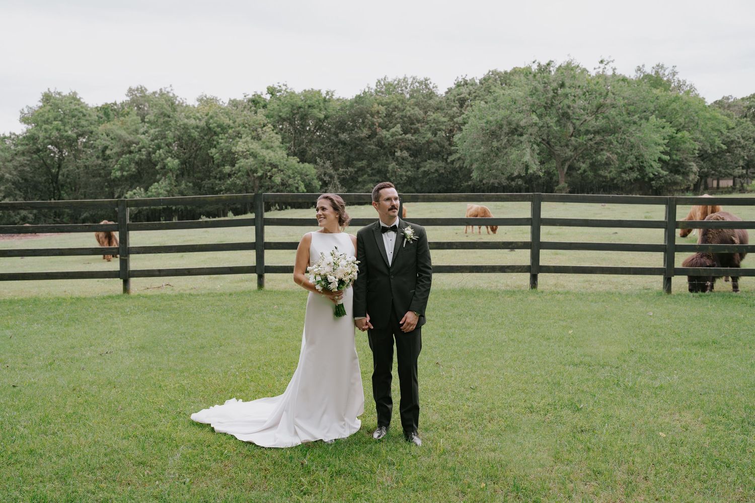 Newlyweds walking together on a green lawn with wooden fencing and trees creating a rustic outdoor setting.