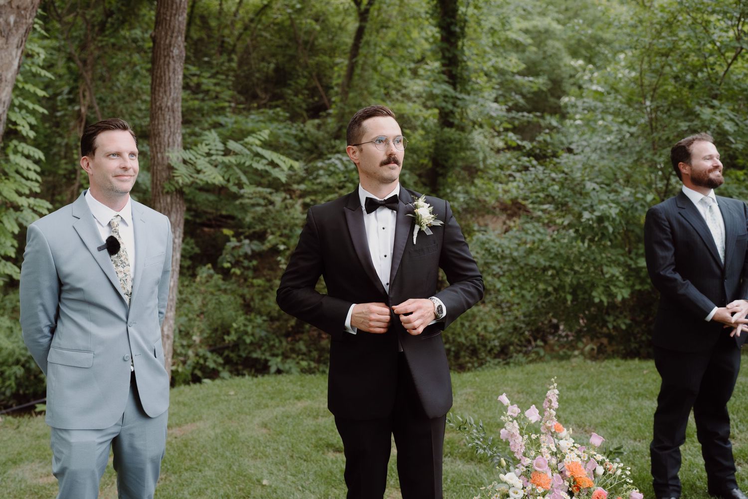 Three groomsmen in formal attire stand at the altar with floral arrangements in a garden setting.