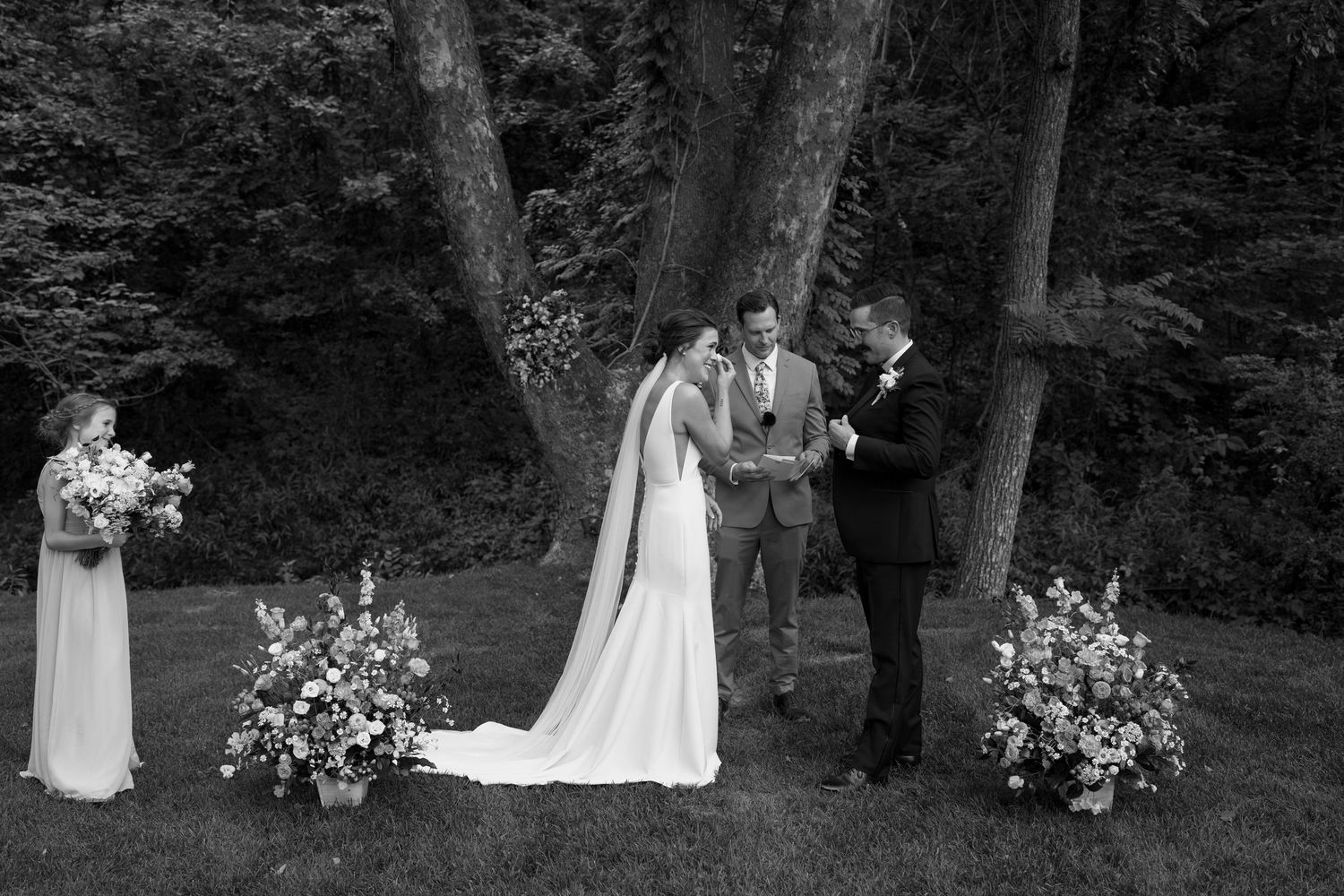 Black and white wedding ceremony photo in a forest setting with floral arrangements.