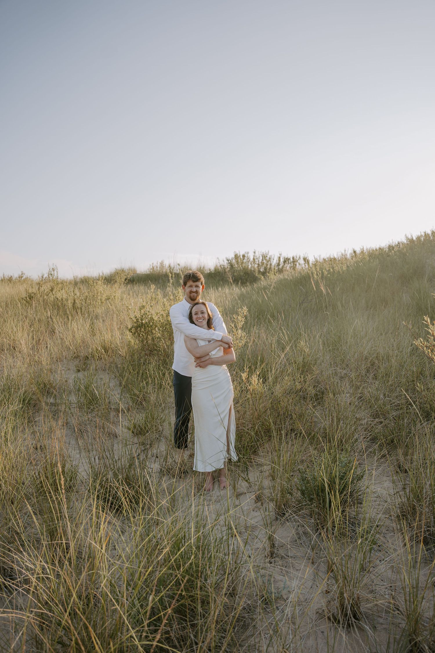 A couple shares an intimate moment among tall grass on a sandy hillside.