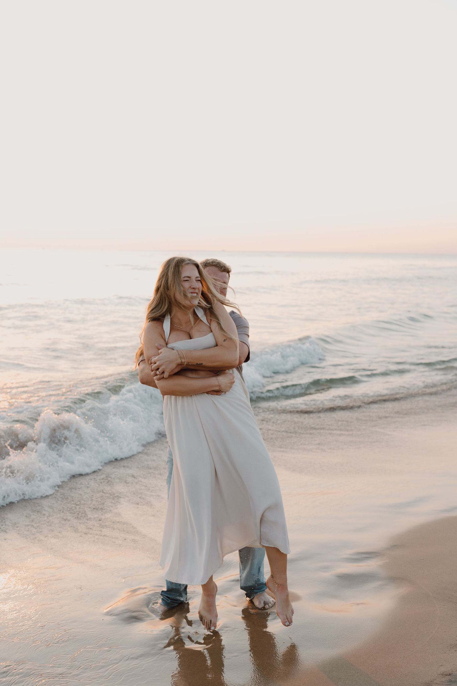 A couple embraces playfully in the shallow ocean waves during sunset on a peaceful beach.