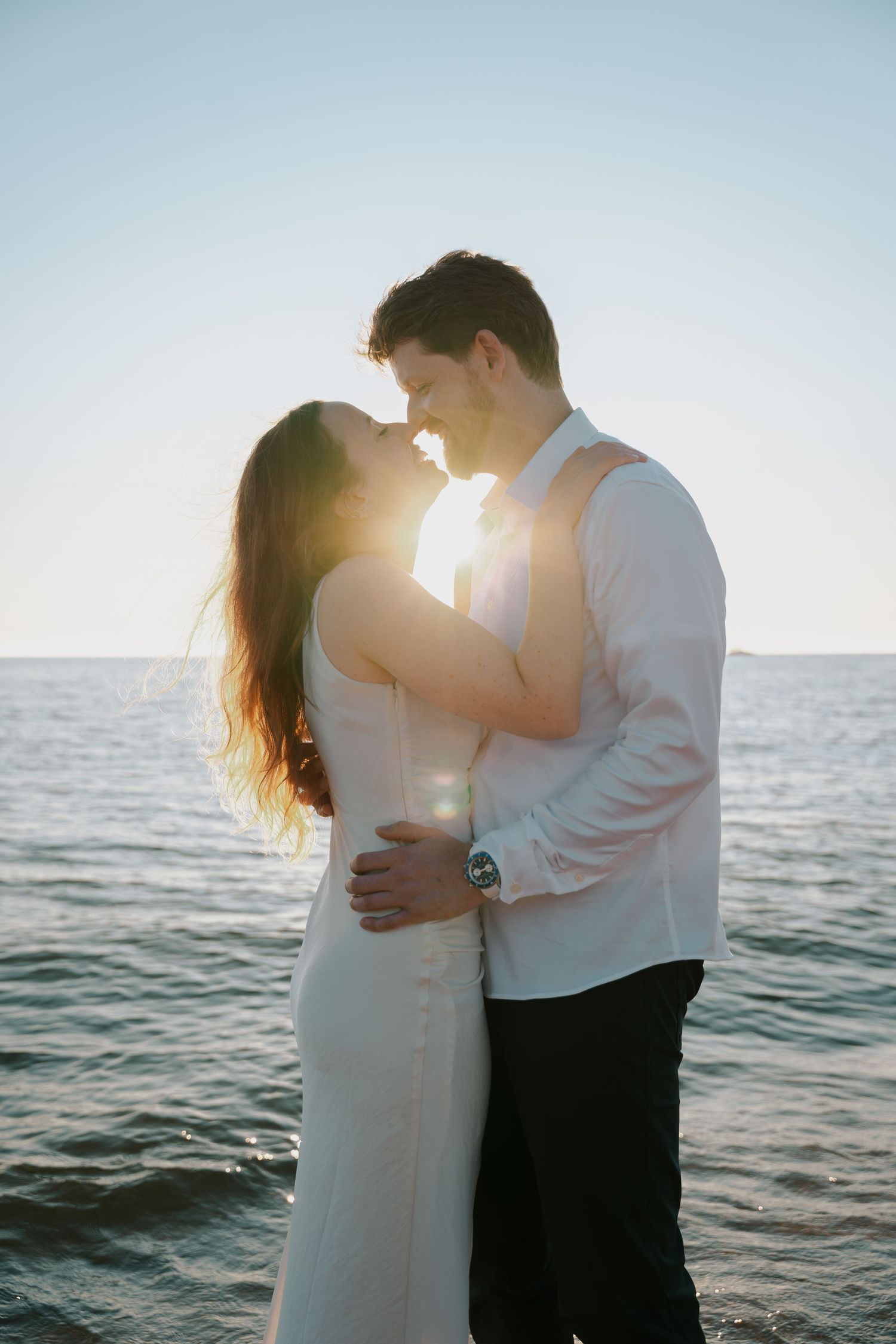 Couple in white and black attire embrace against serene ocean backdrop.