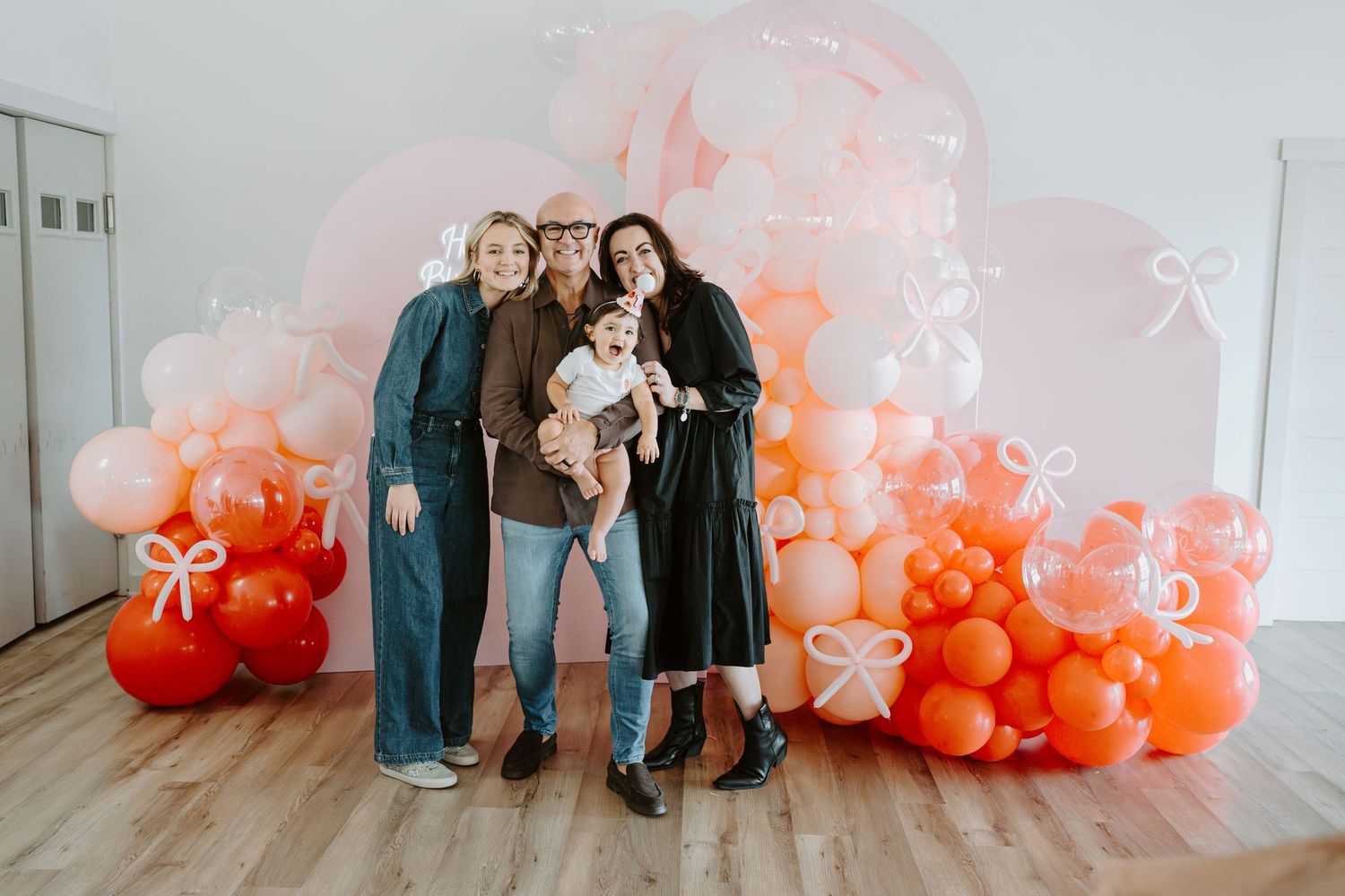 Birthday party setup with coral and peach balloon garland against white wall with wooden flooring.