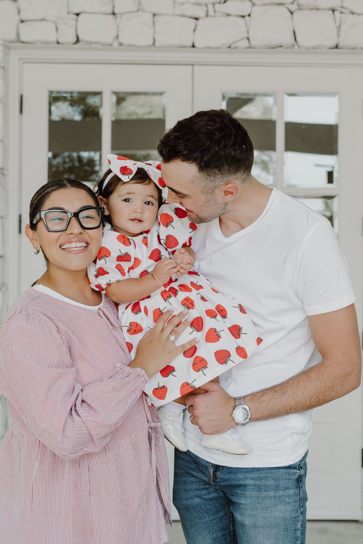 A sweet family embrace in front of a white stone wall with glass doors.