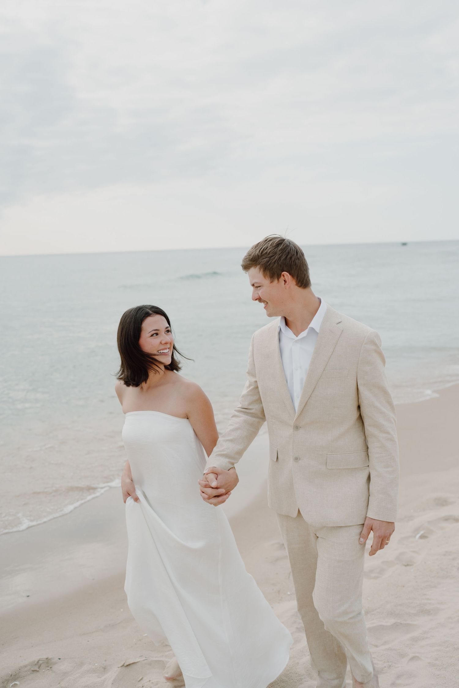 A couple in white and beige attire shares a romantic moment while walking along a serene beach.