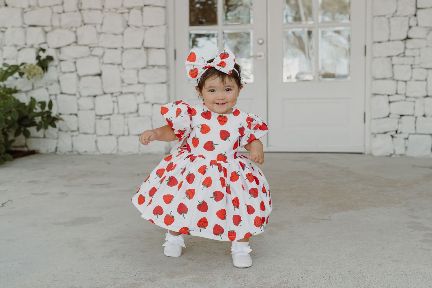Toddler in polka dot dress and bow twirls on white porch.