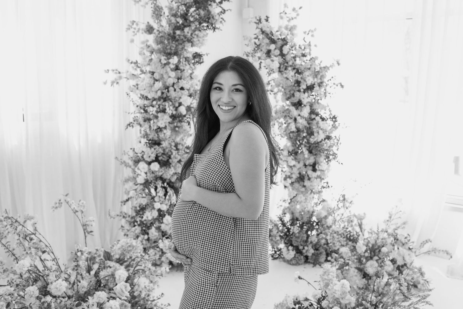 Maternity photoshoot featuring a checkered dress against a backdrop of pink and peach floral arrangements.