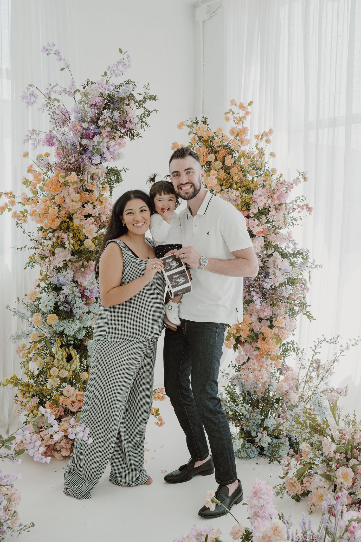 Family photo session with floral backdrop featuring pink and peach blooms while holding ultrasound image.