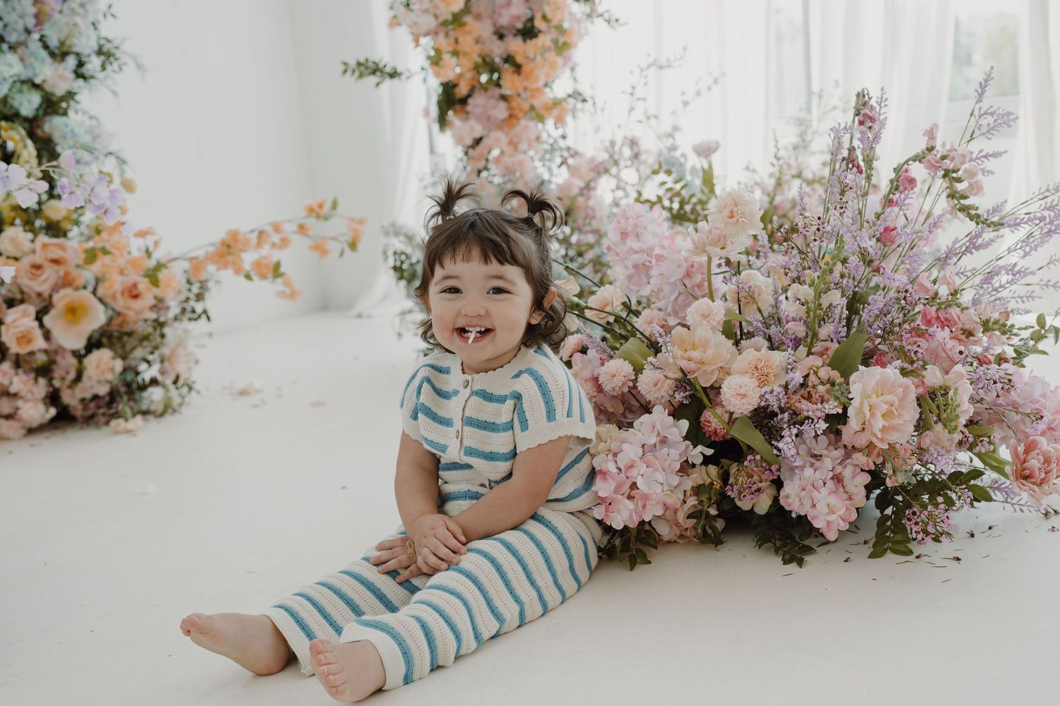 Child in blue striped outfit sitting beside pink and peach floral arrangements.