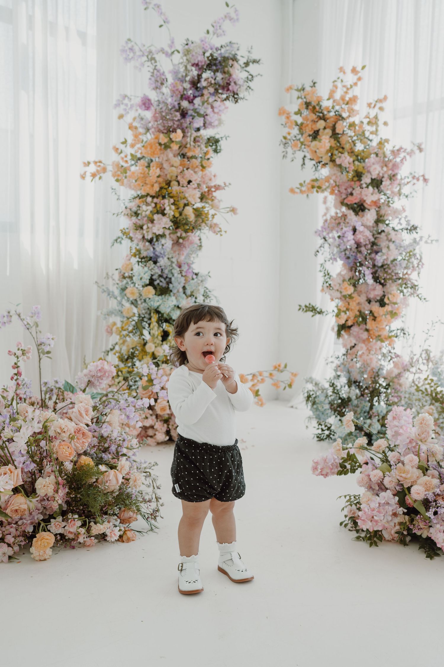 Joyful toddler playing in bright studio with pastel floral arrangements.