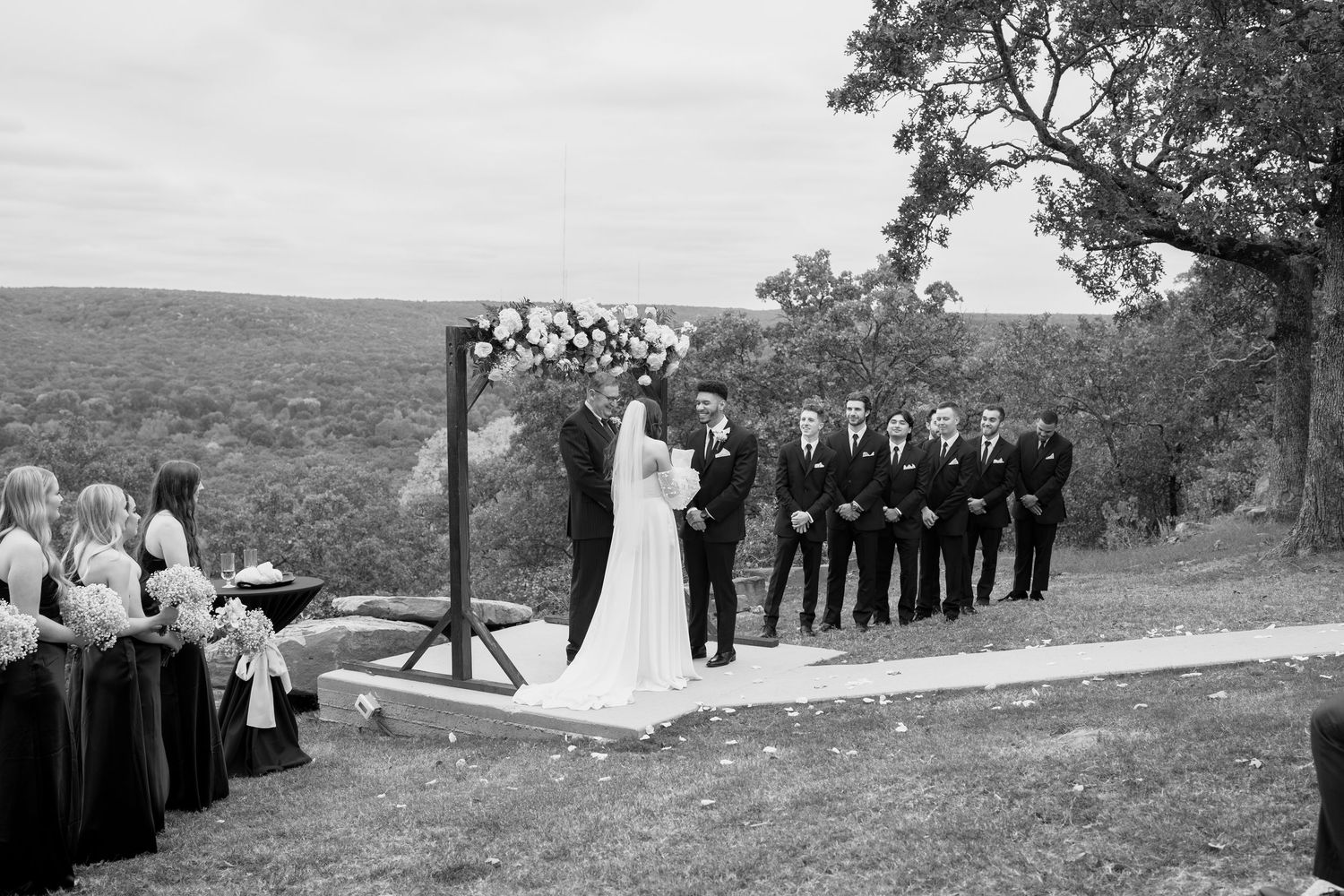 Black and white wedding photo showing ceremony setup with floral arch and wedding party on hillside.