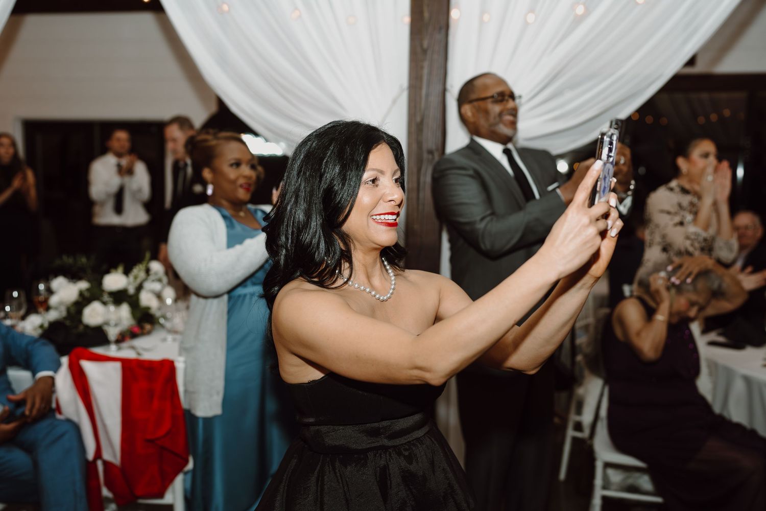 Guest in black dress taking photos during wedding reception with white drapes in background.