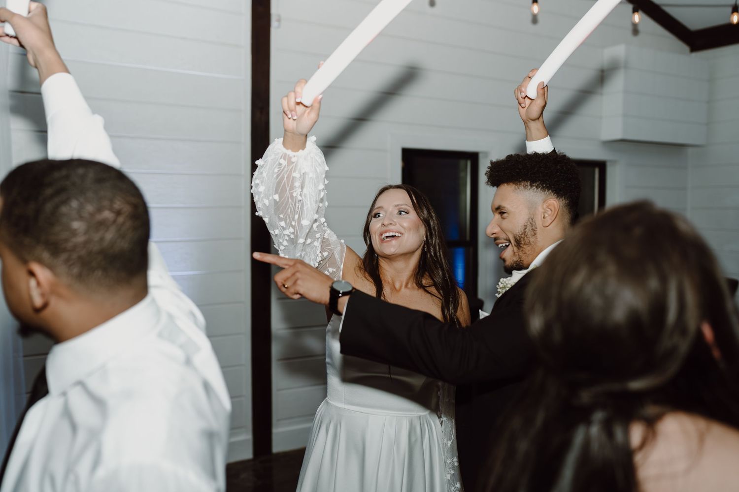 Party guests wave LED foam sticks while dancing at an indoor wedding reception.