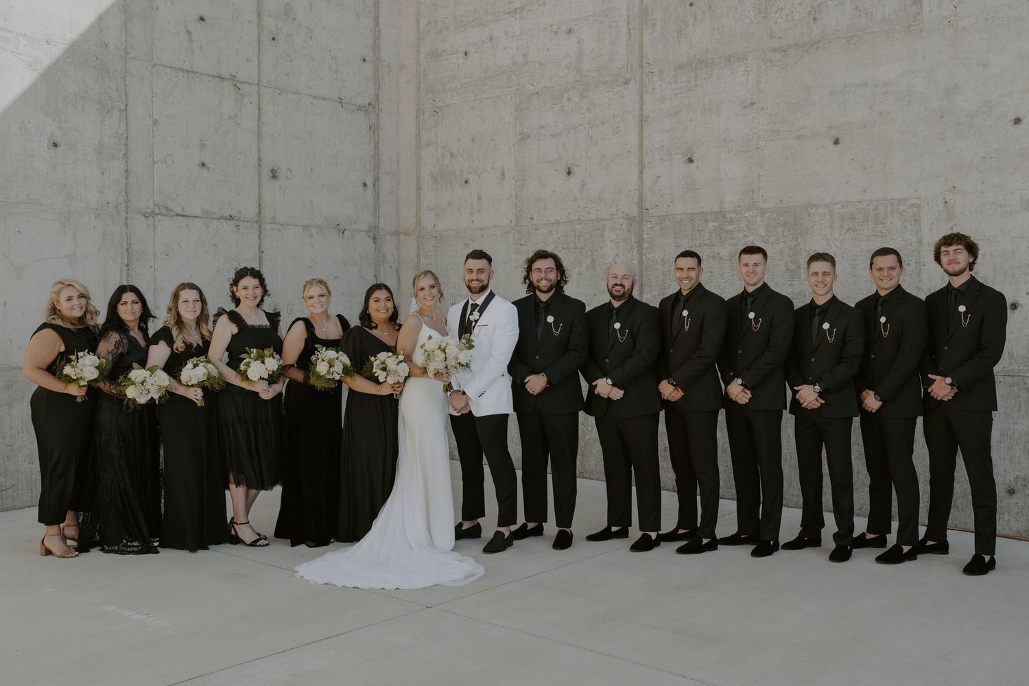 A wedding party poses against a grey wall in formal black and white attire for group photos.