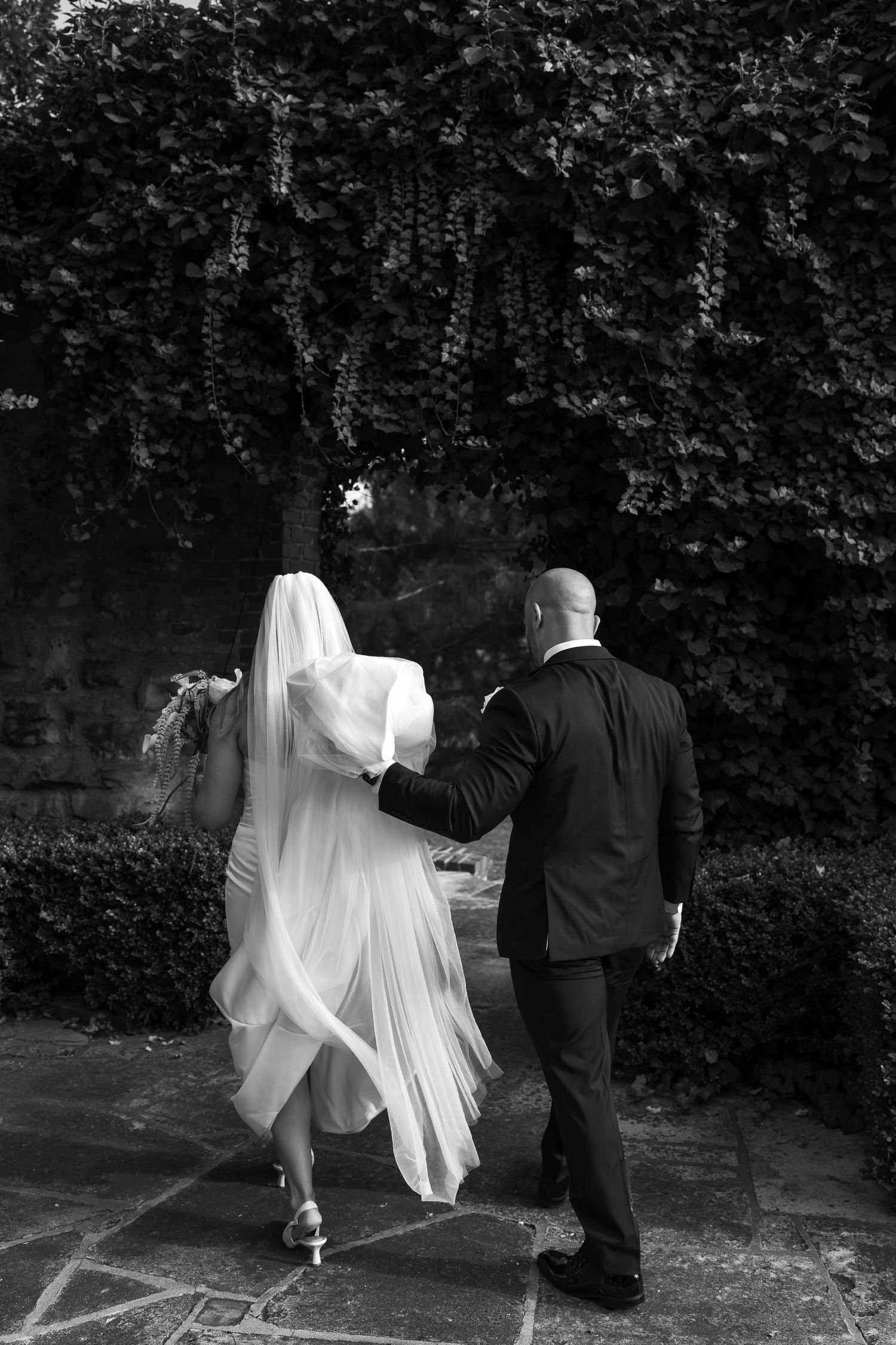 Black and white photo of bride and groom walking hand in hand through a garden with ivy-covered walls.