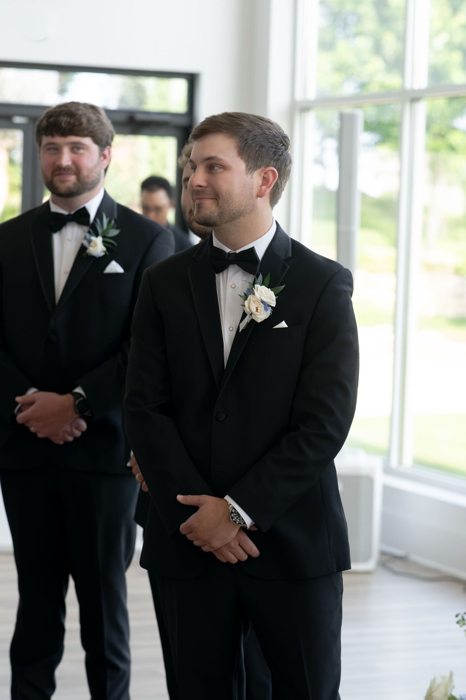 Groom in black tuxedo with white boutonniere smiling at altar, groomsmen standing behind him at wedding ceremony.