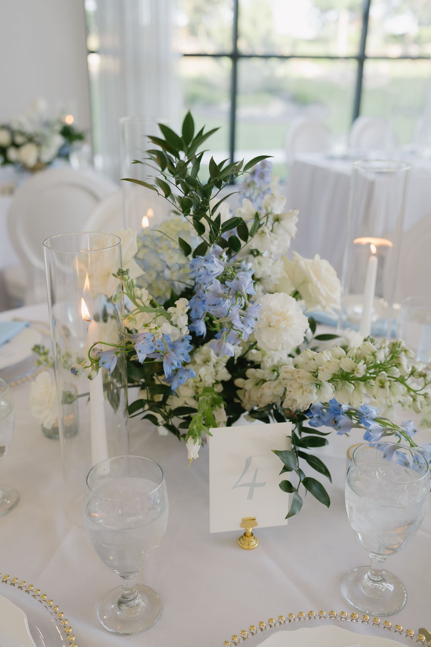 Elegant wedding table centerpiece with blue and white flowers, greenery, candles, and a gold-rimmed table number card.