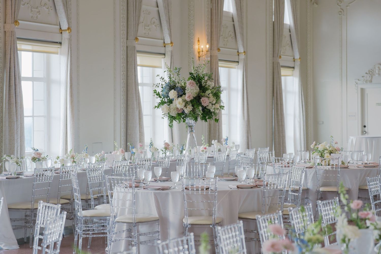 Elegant wedding reception hall with ghost chairs, round tables, white linens, and tall floral centerpieces in a bright ballroom.