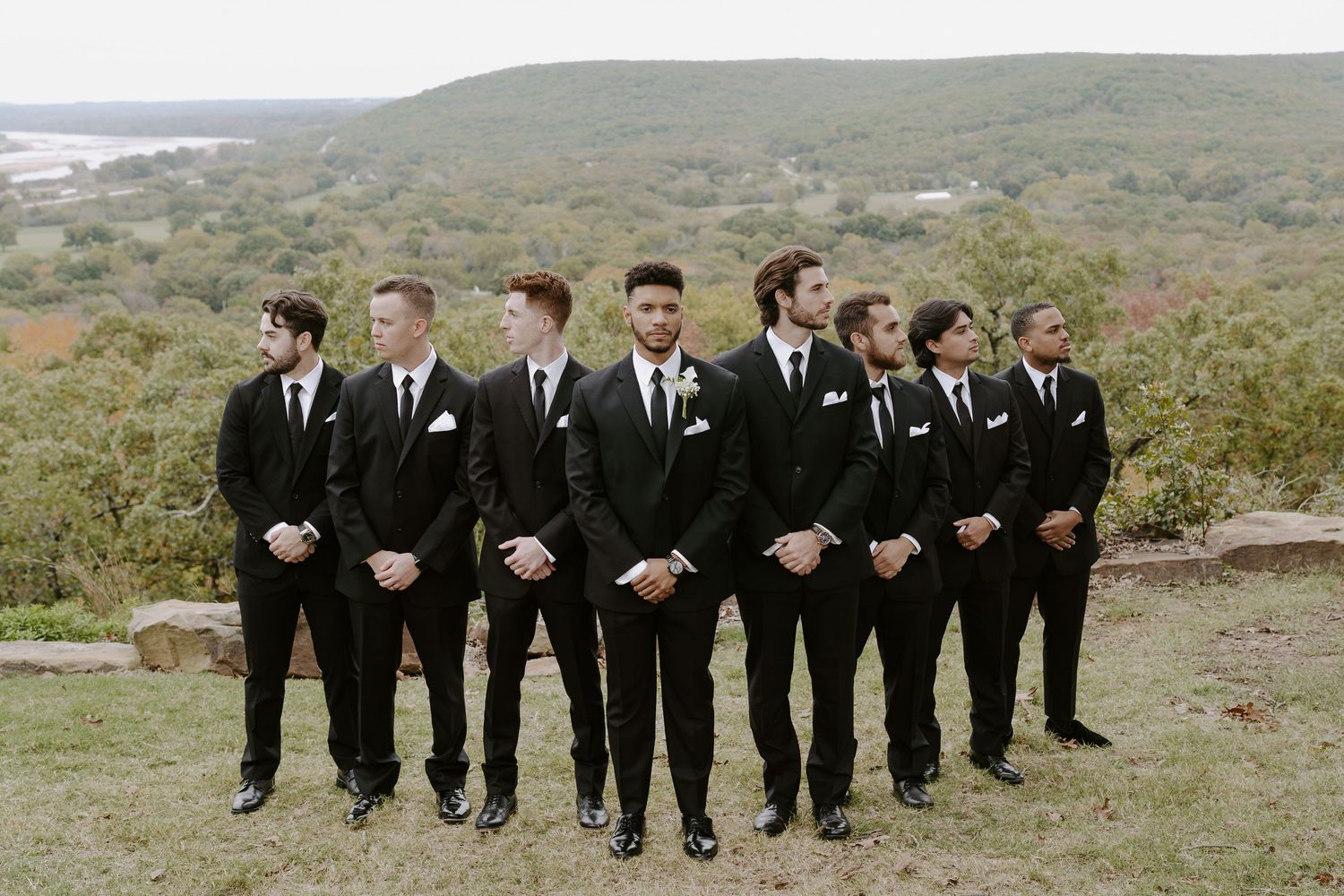 Wedding party lined up in black suits and ties against rolling hills landscape.