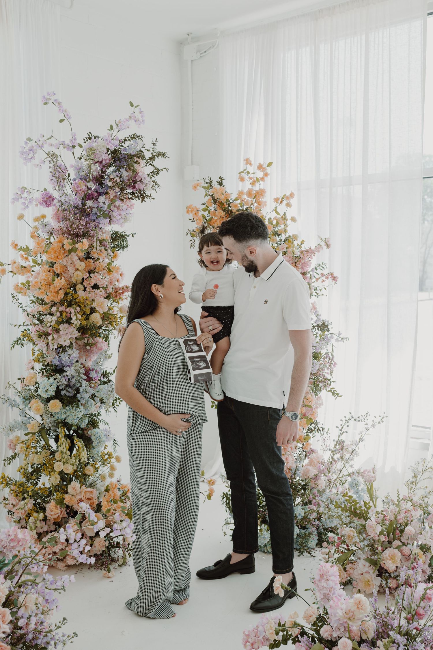 A family celebrates a pregnancy announcement with ultrasound photos in front of blooming flowers.