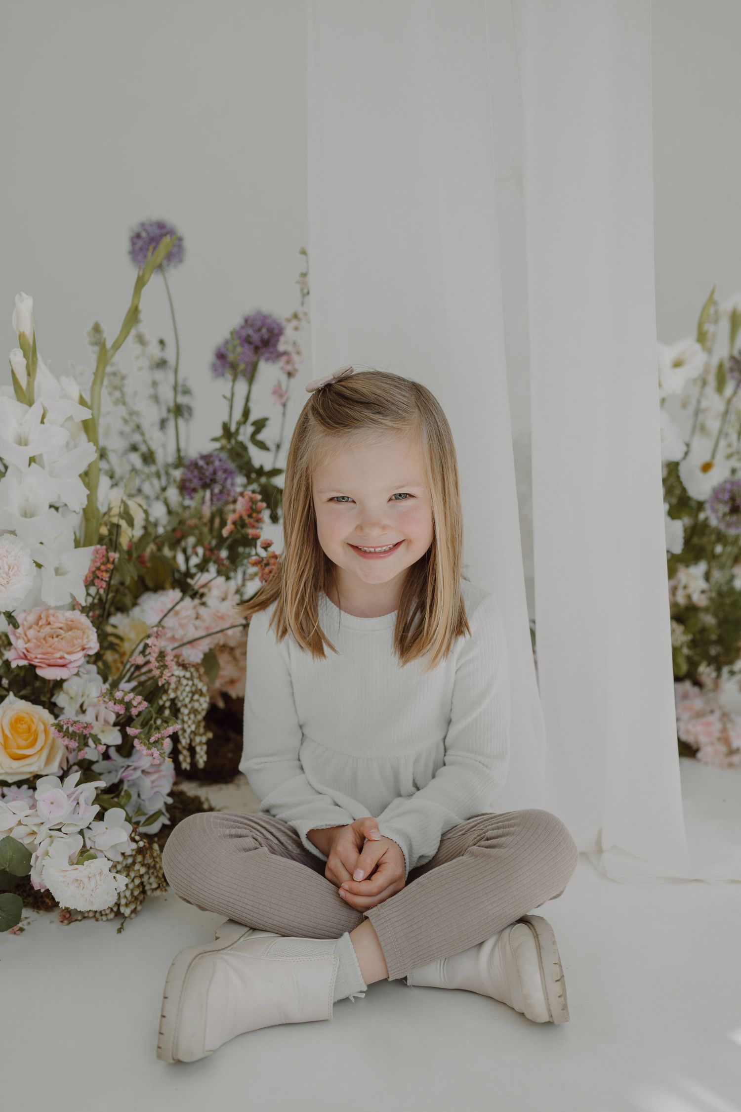 Portrait in white room with floral arrangements featuring soft pink roses and purple blooms against white curtains.
