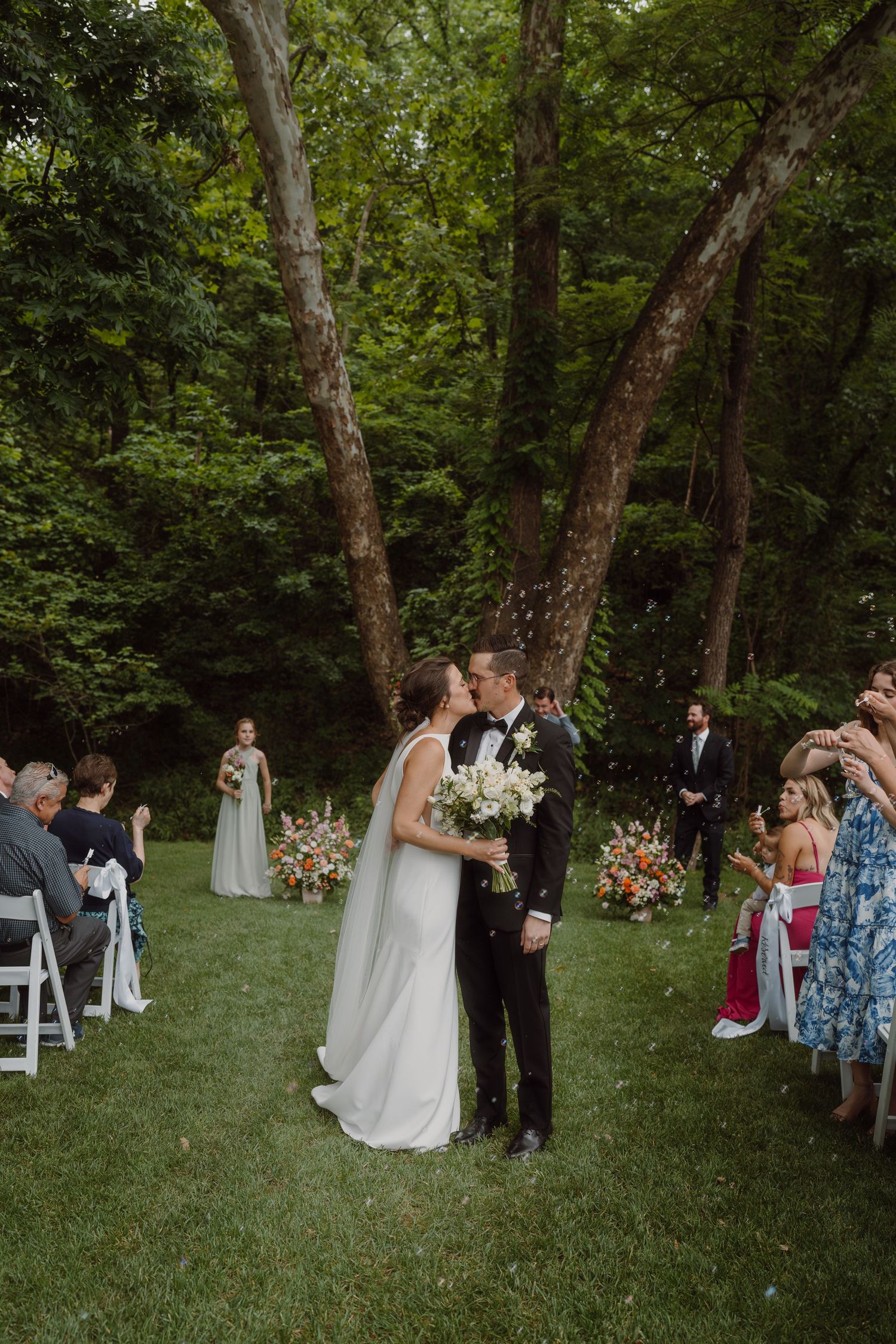 An intimate forest wedding ceremony with white chairs and floral decorations among tall trees.