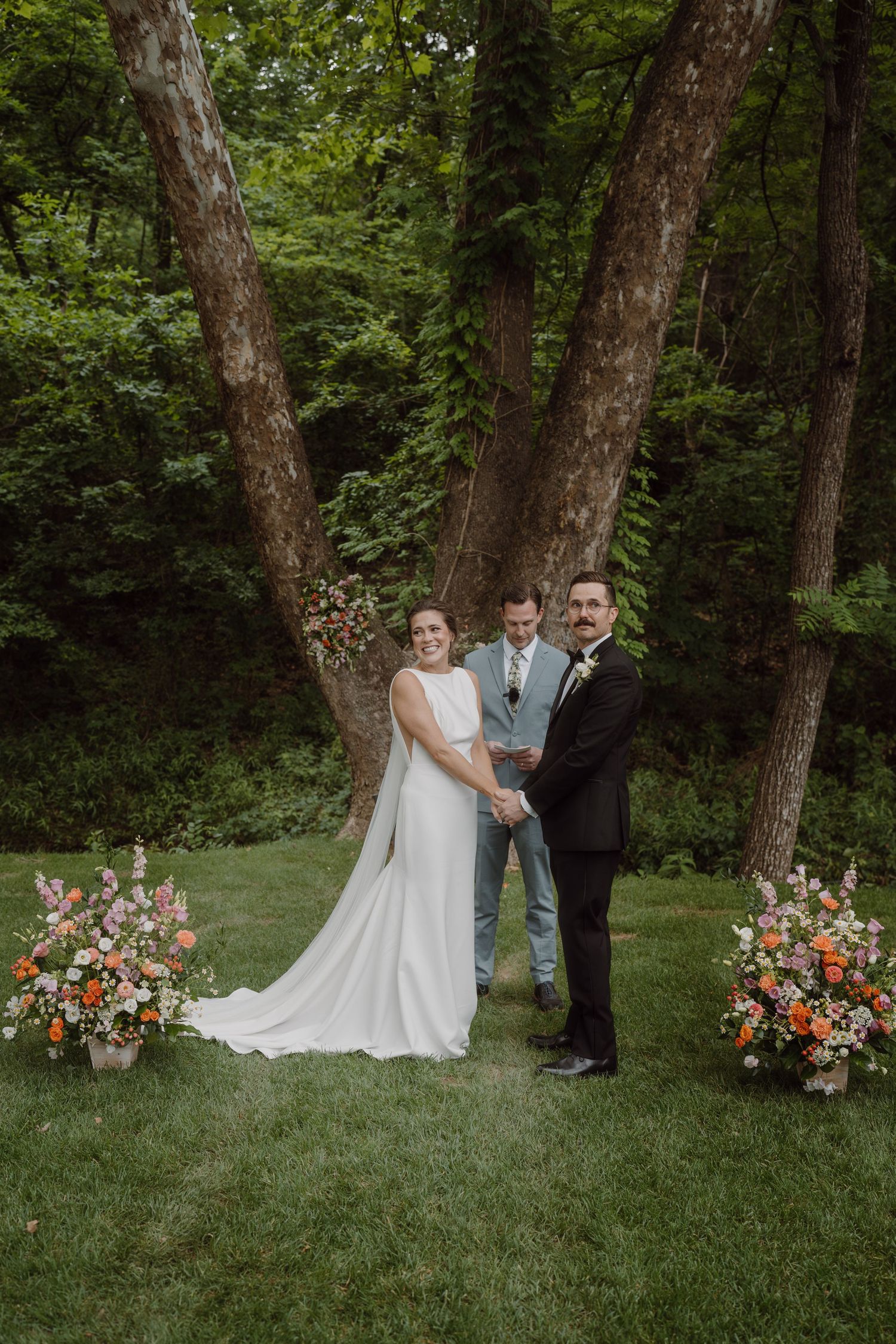 Wedding ceremony participants gather under tall trees with colorful floral arrangements.