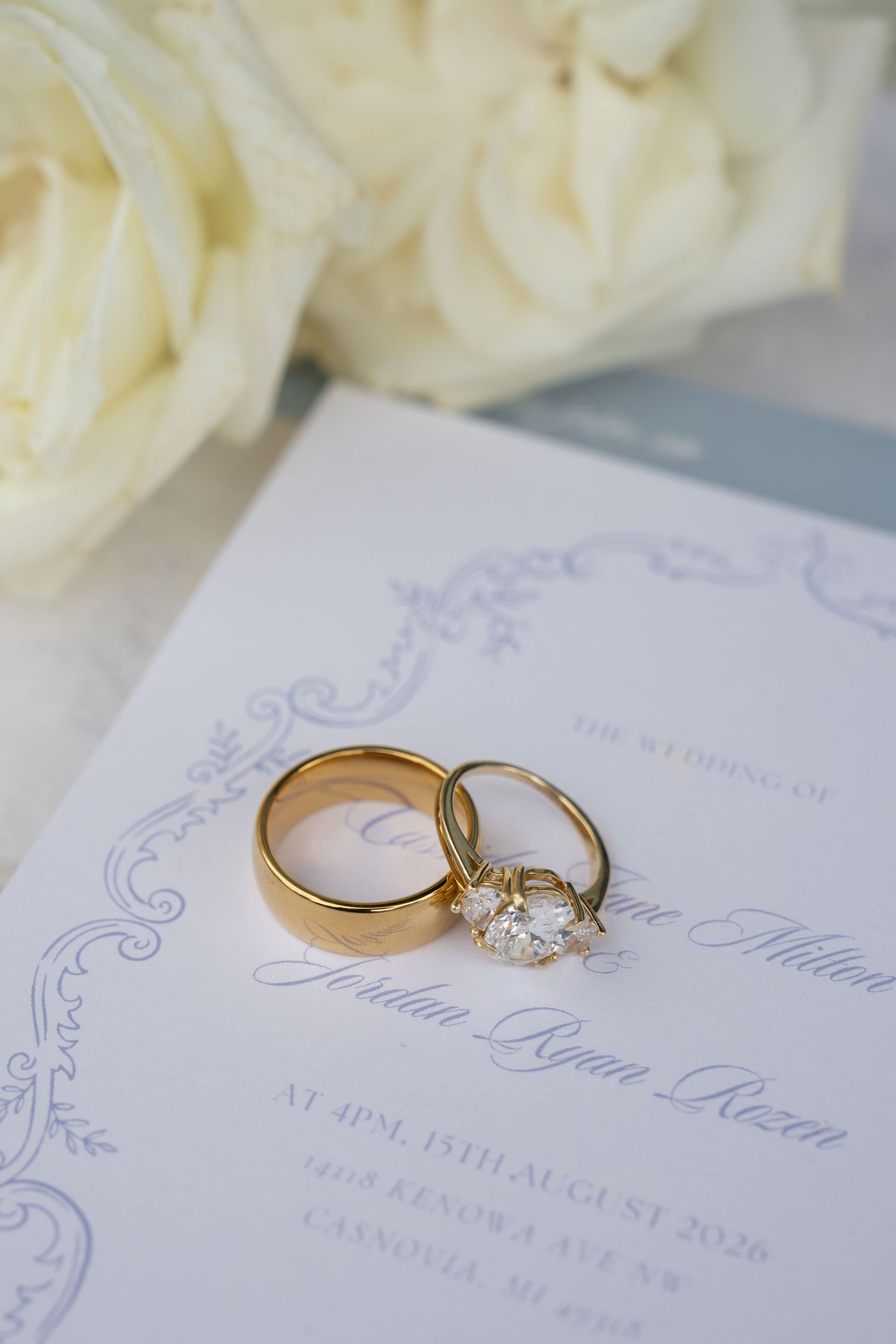 Gold wedding bands and diamond engagement ring rest on elegant wedding invitation beside white roses.