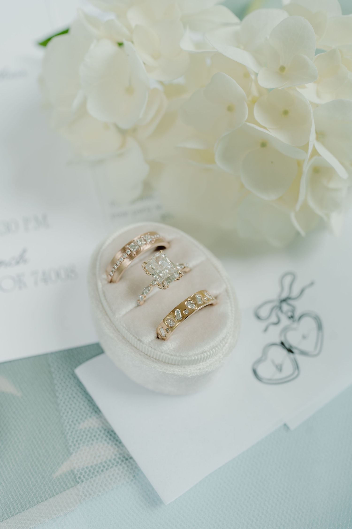 Three diamond wedding rings displayed in a velvet ring box beside white hydrangeas and elegant stationery.