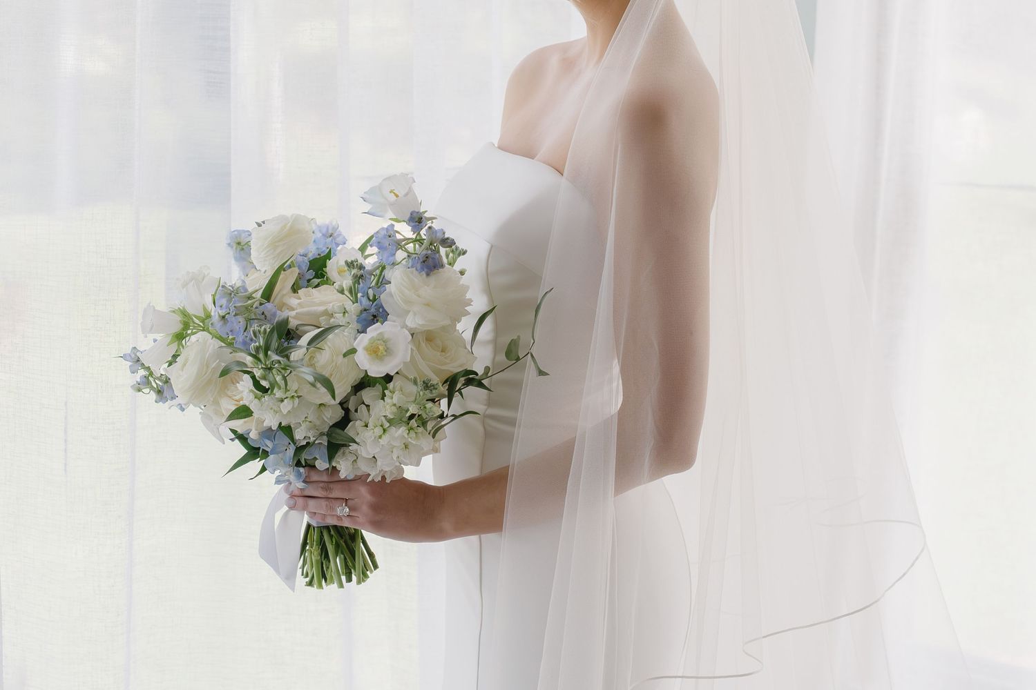 Bride in white strapless gown holding elegant blue and white floral wedding bouquet with veil draped softly.