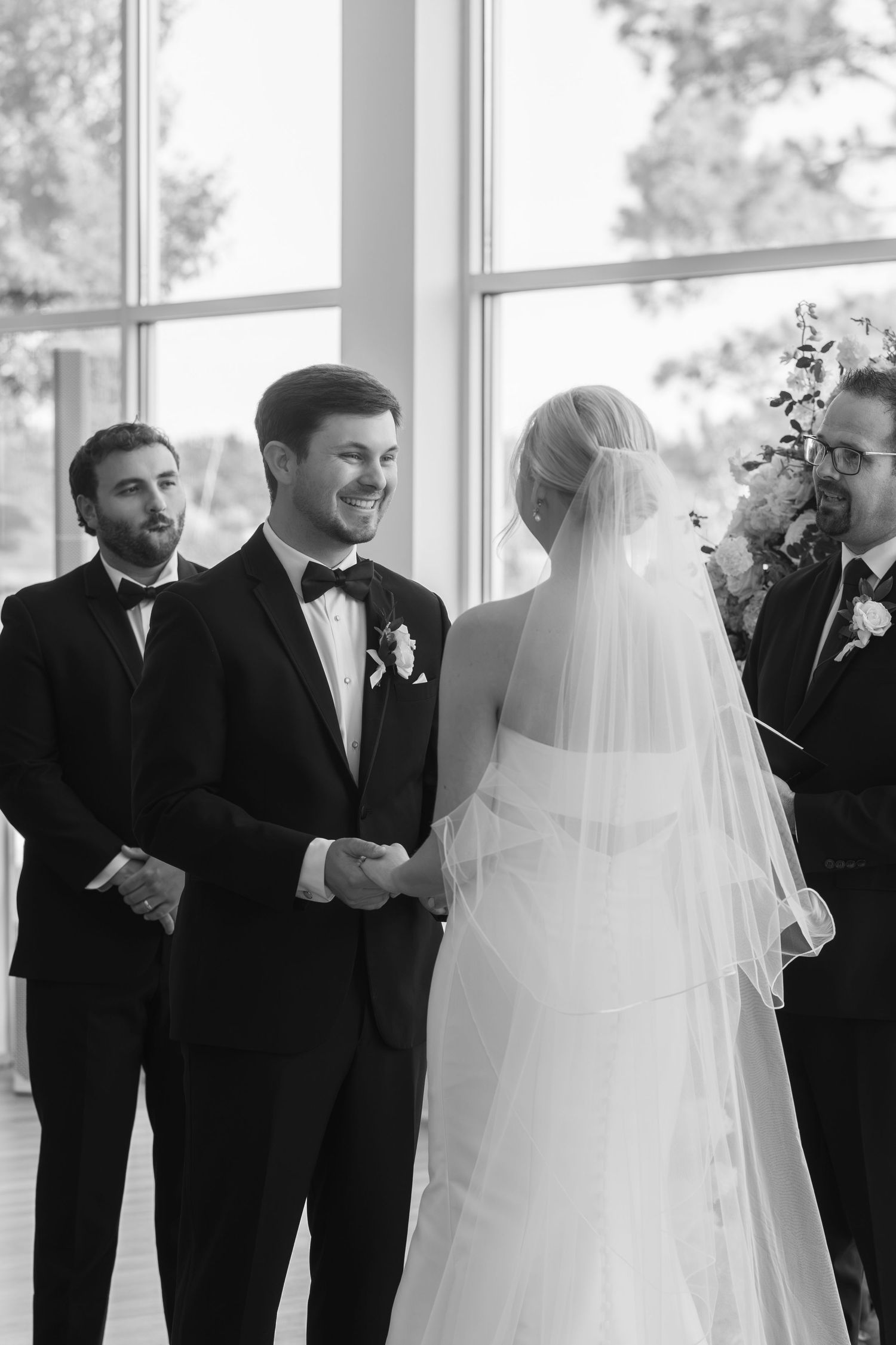 Black and white wedding ceremony photo of smiling groom holding bride's hands at the altar.