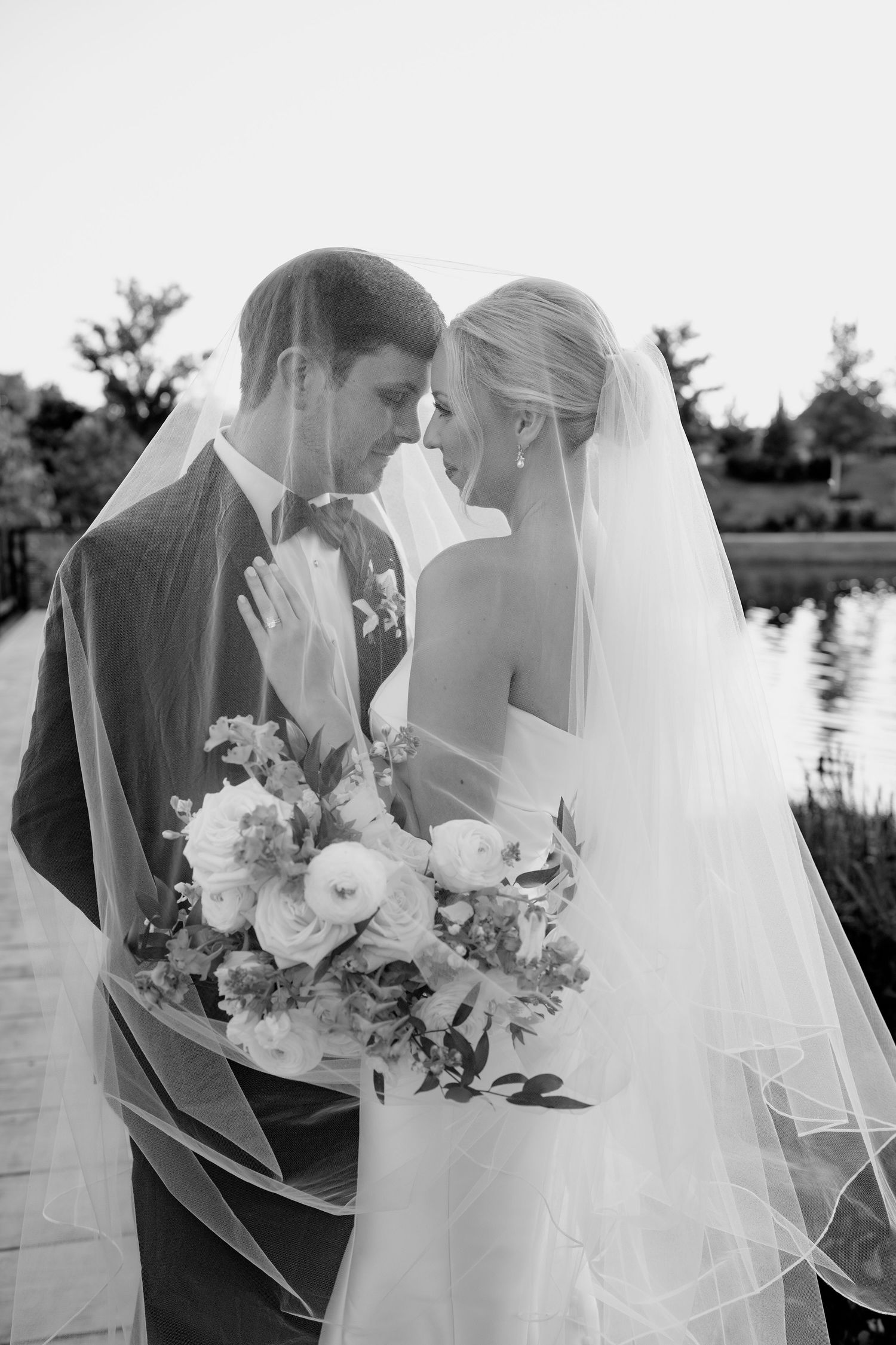 Black and white photo of bride and groom sharing an intimate moment under veil, bride holding bouquet near a lake.