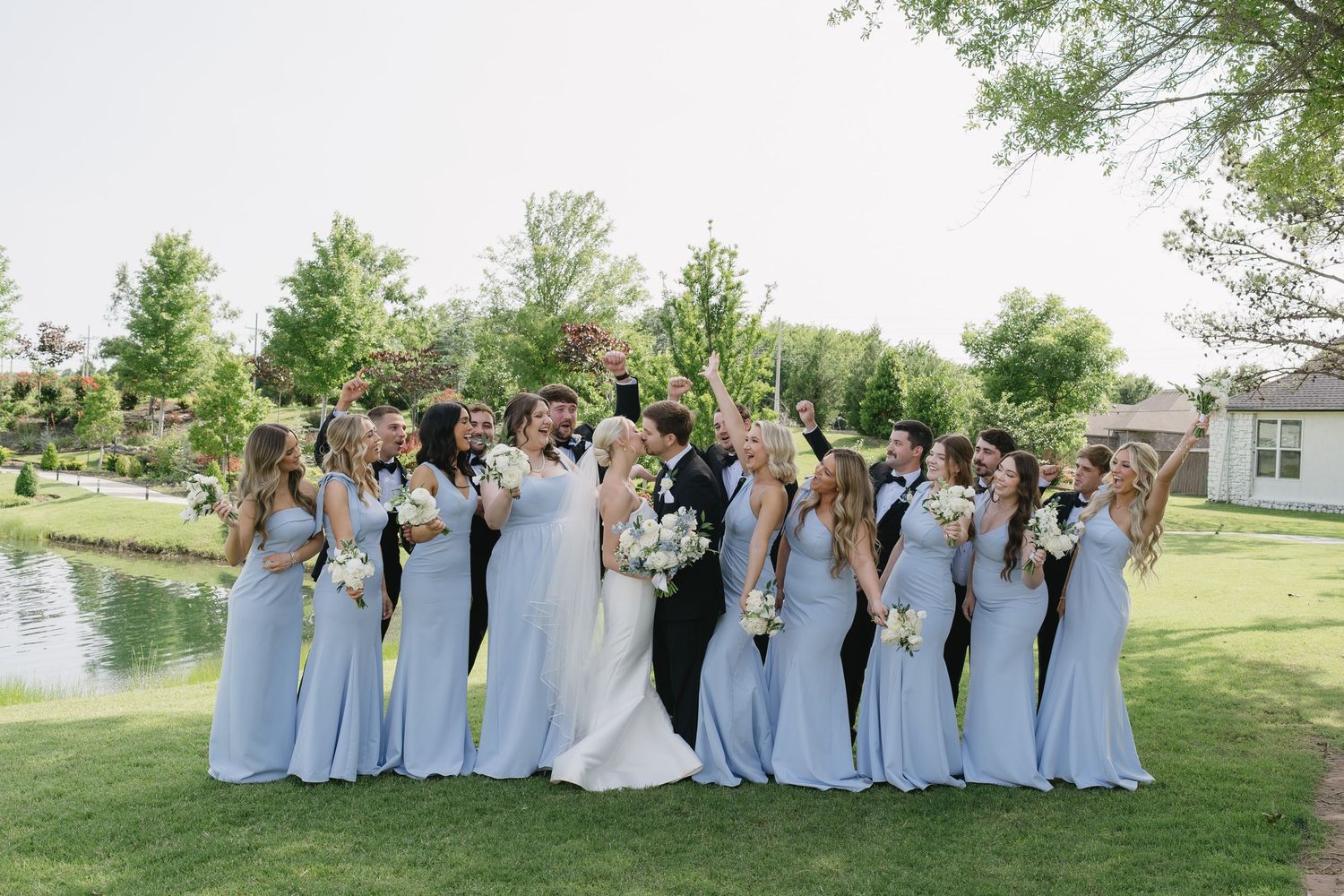 Bride and groom kiss surrounded by bridesmaids in light blue gowns holding white bouquets on a lush green lawn.