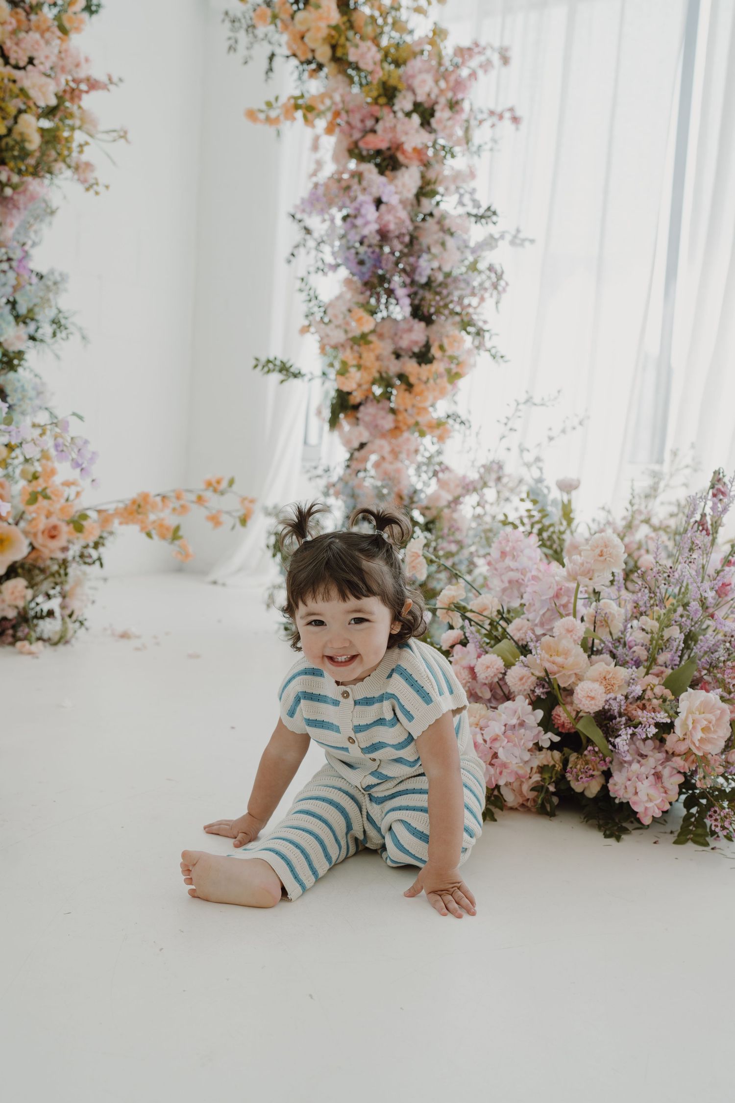 Playful toddler in striped romper posing by elegant floral backdrop.