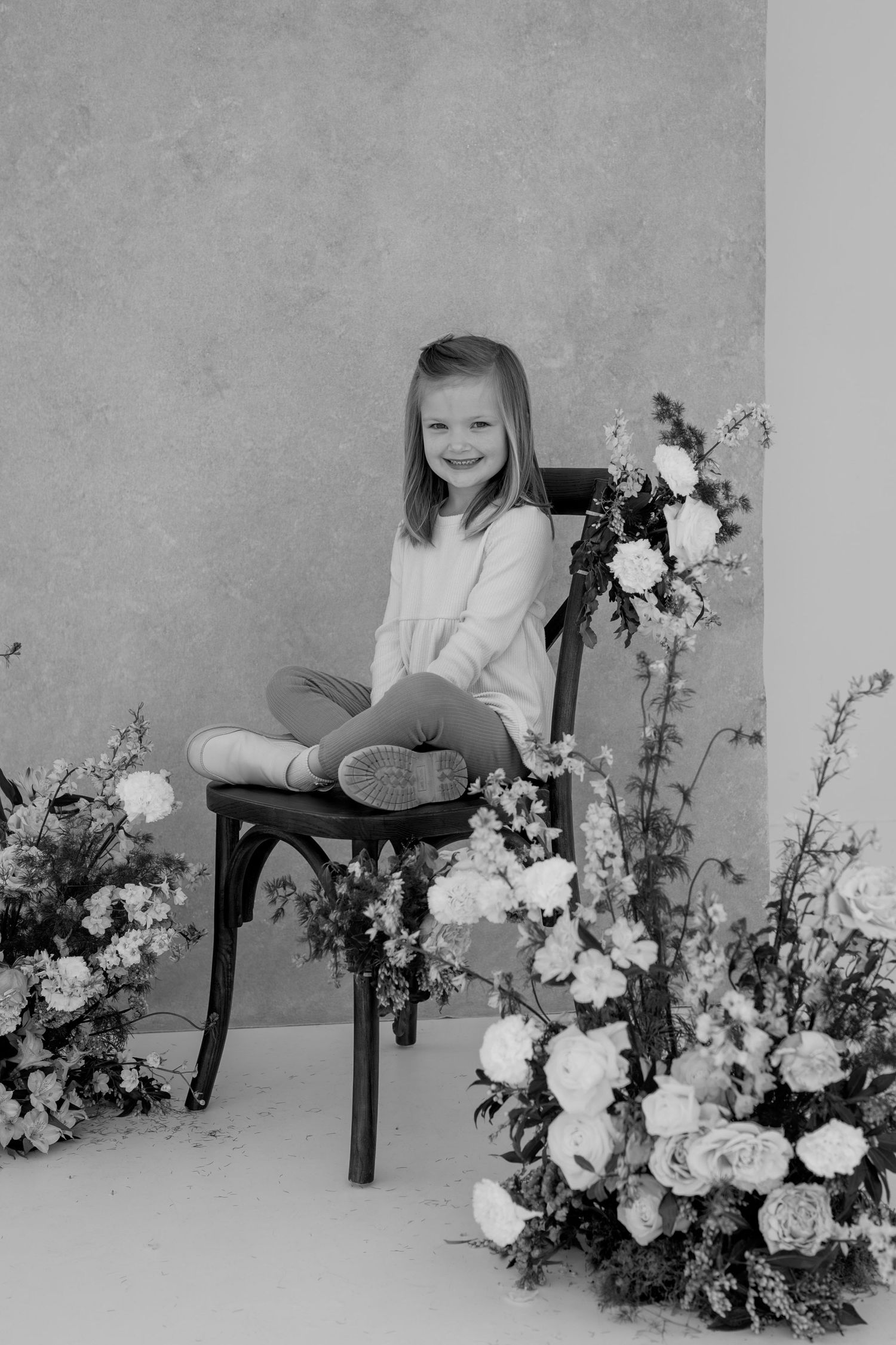 Black and white studio portrait with floral decorations and dark wooden chair.