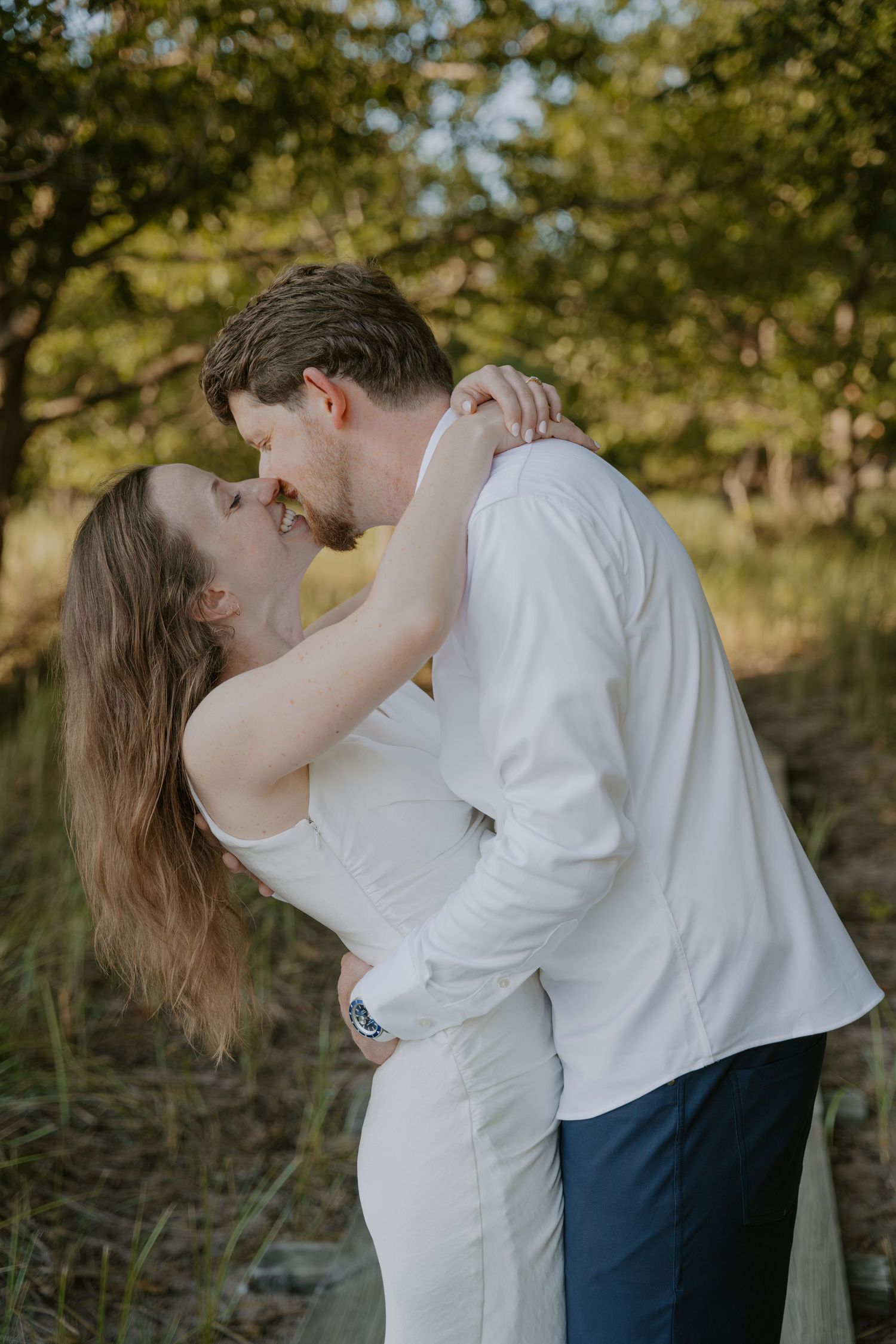 A romantic couple embraces and kisses in a natural outdoor setting with golden sunlight filtering through trees.