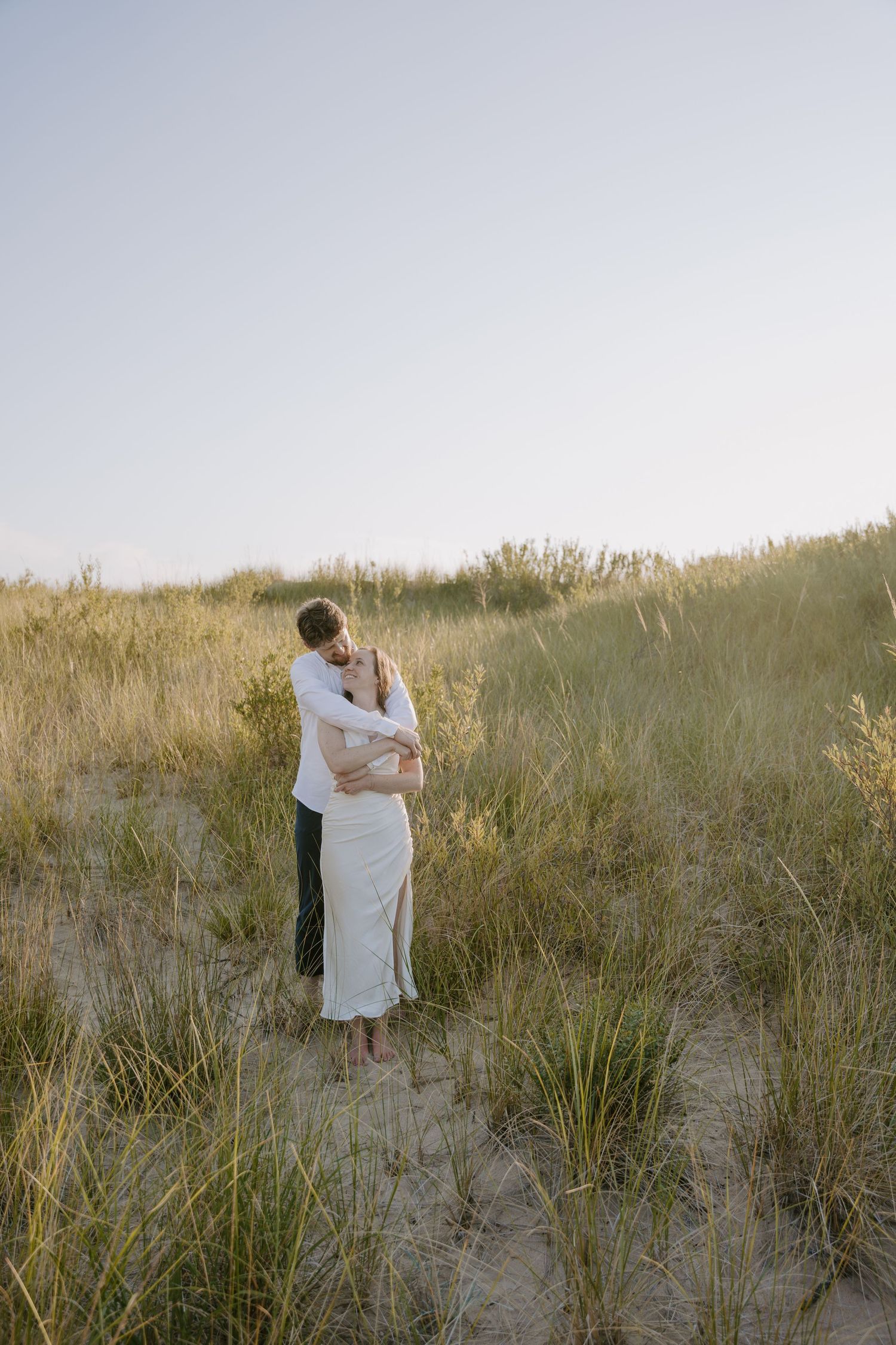 Two figures wrapped in white fabric embrace on coastal dunes at sunset.