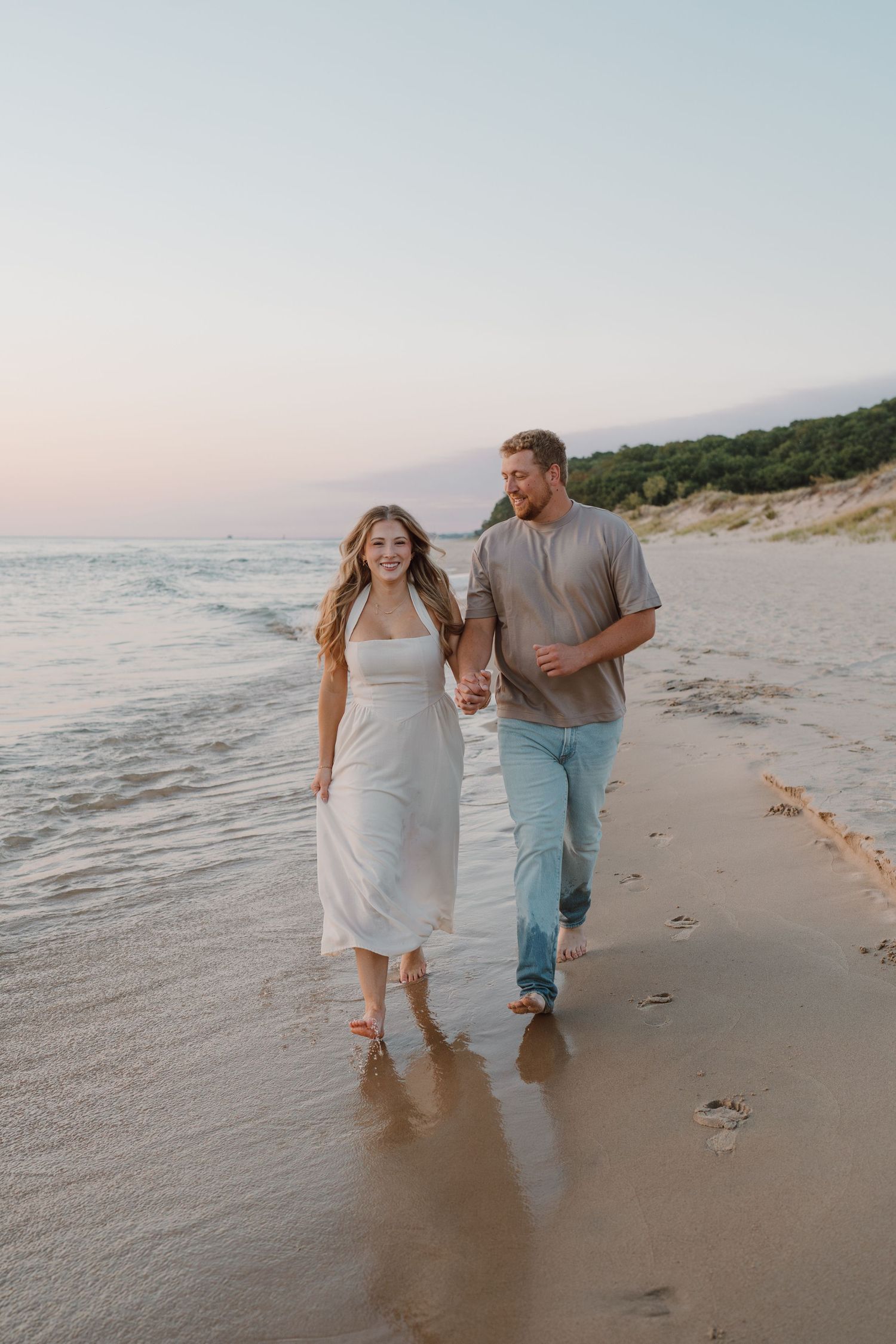 Couple walking hand in hand along shoreline during golden hour.