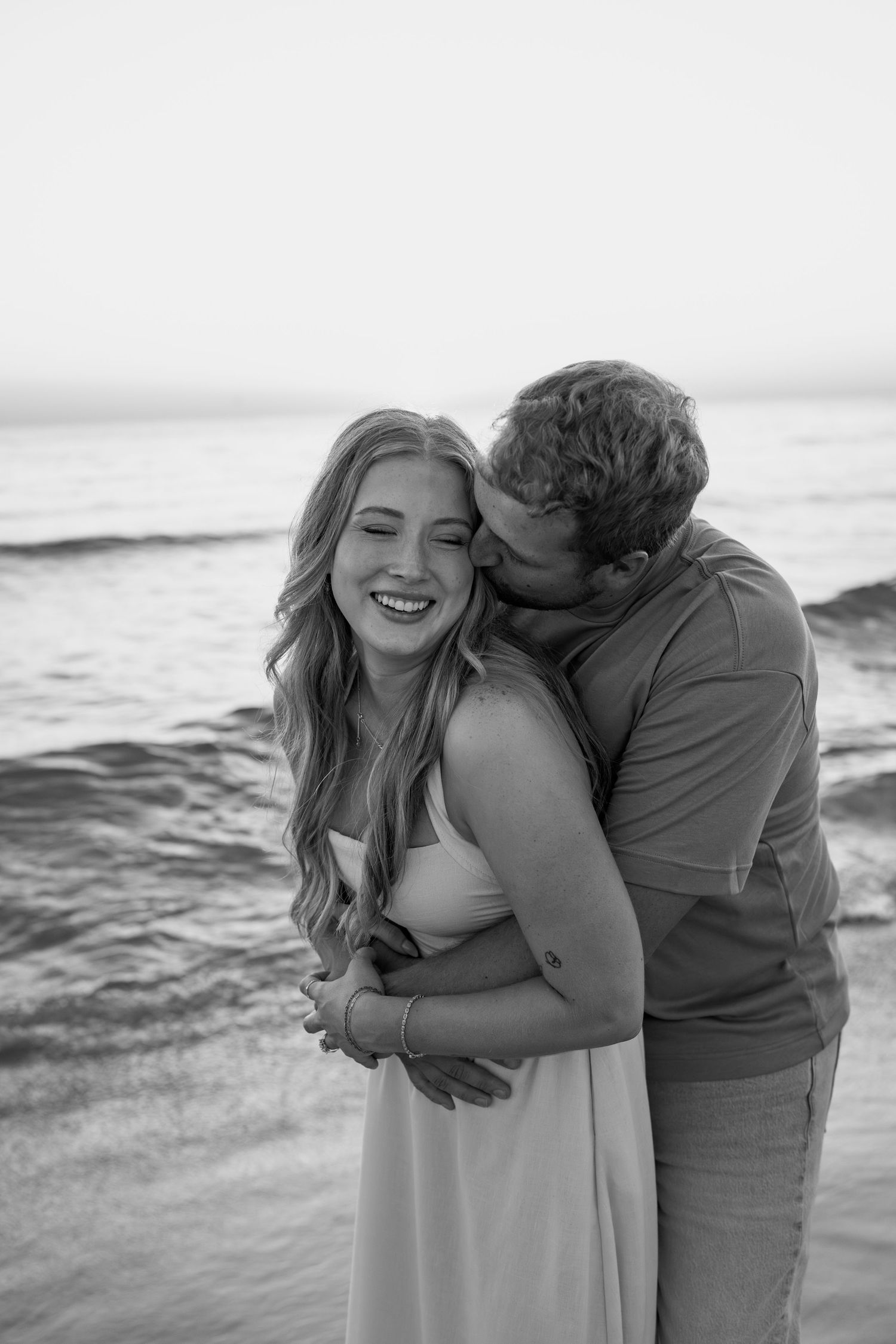 A couple shares intimate moments and laughter on a beach with waves crashing behind them.