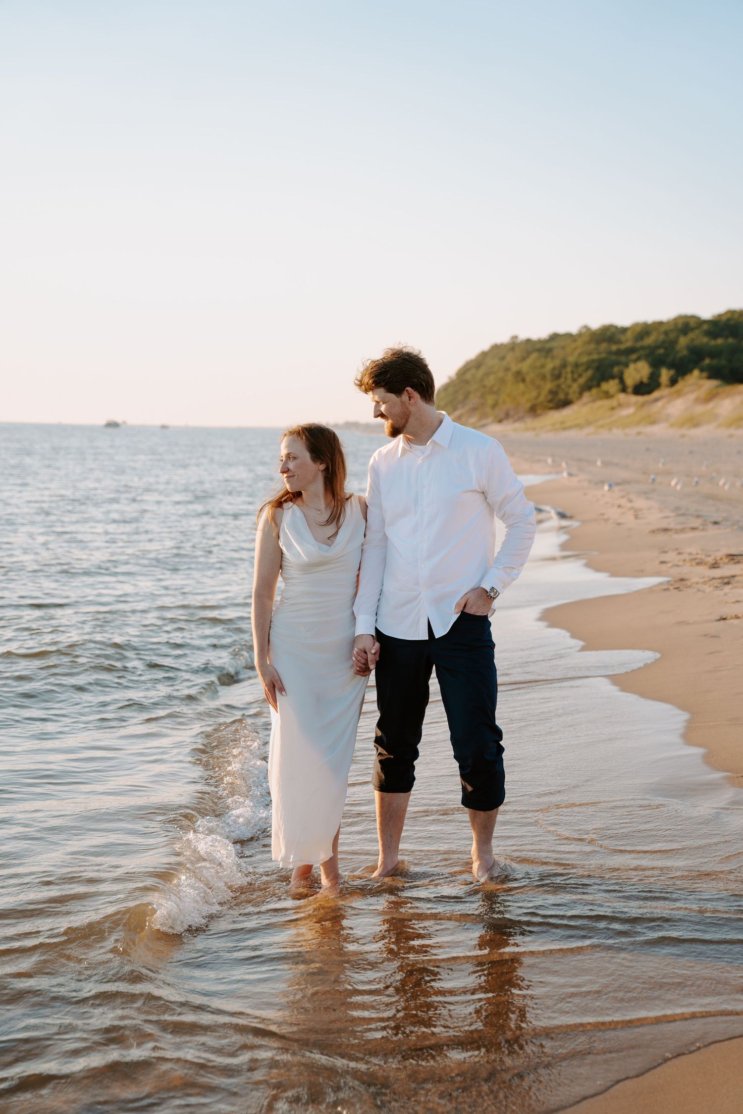 A couple walks together along the beach shoreline at sunset, their feet in the gentle waves.