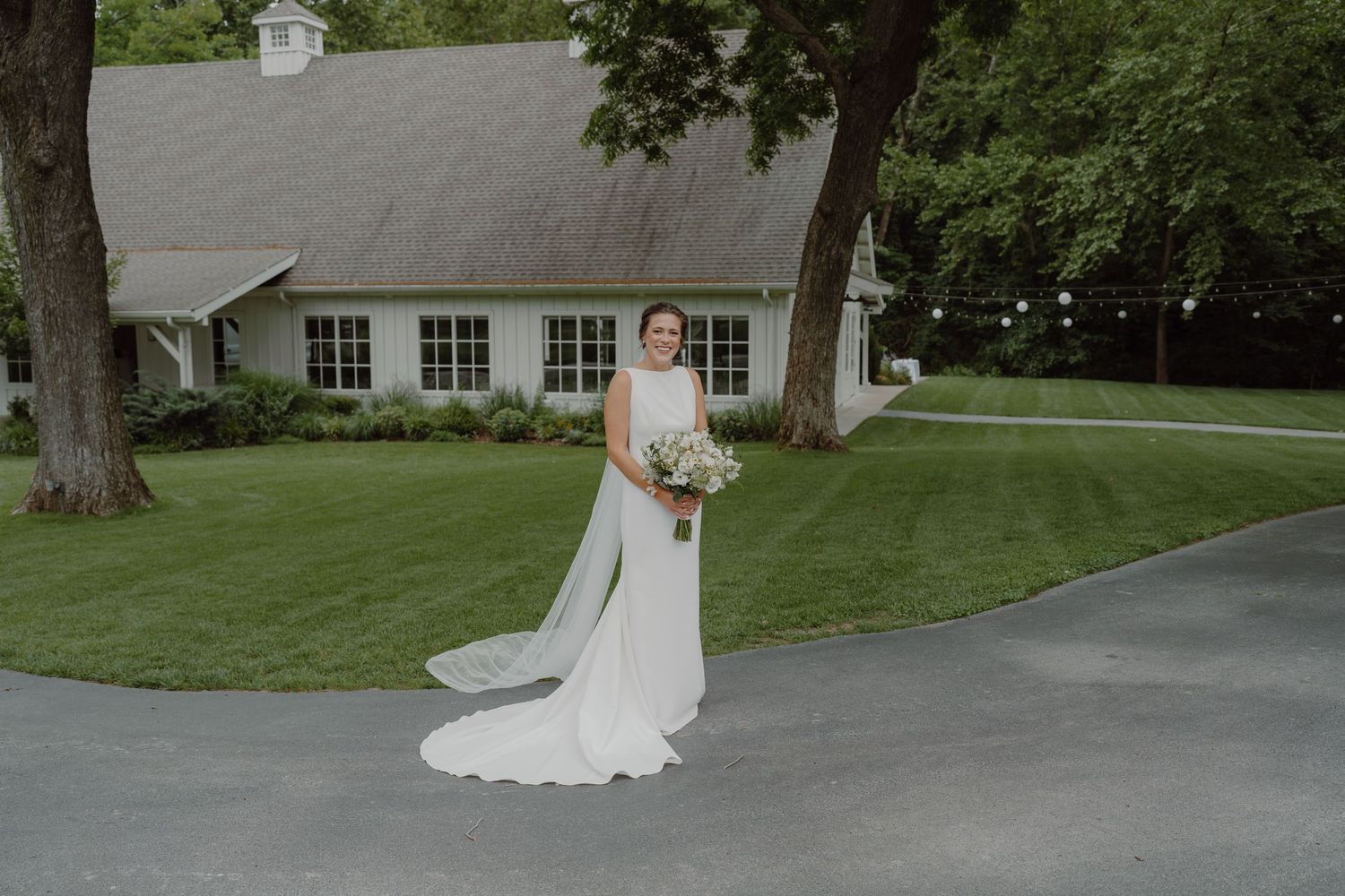 A bride in a sleeveless white gown stands on a paved path near a white building with multiple windows.