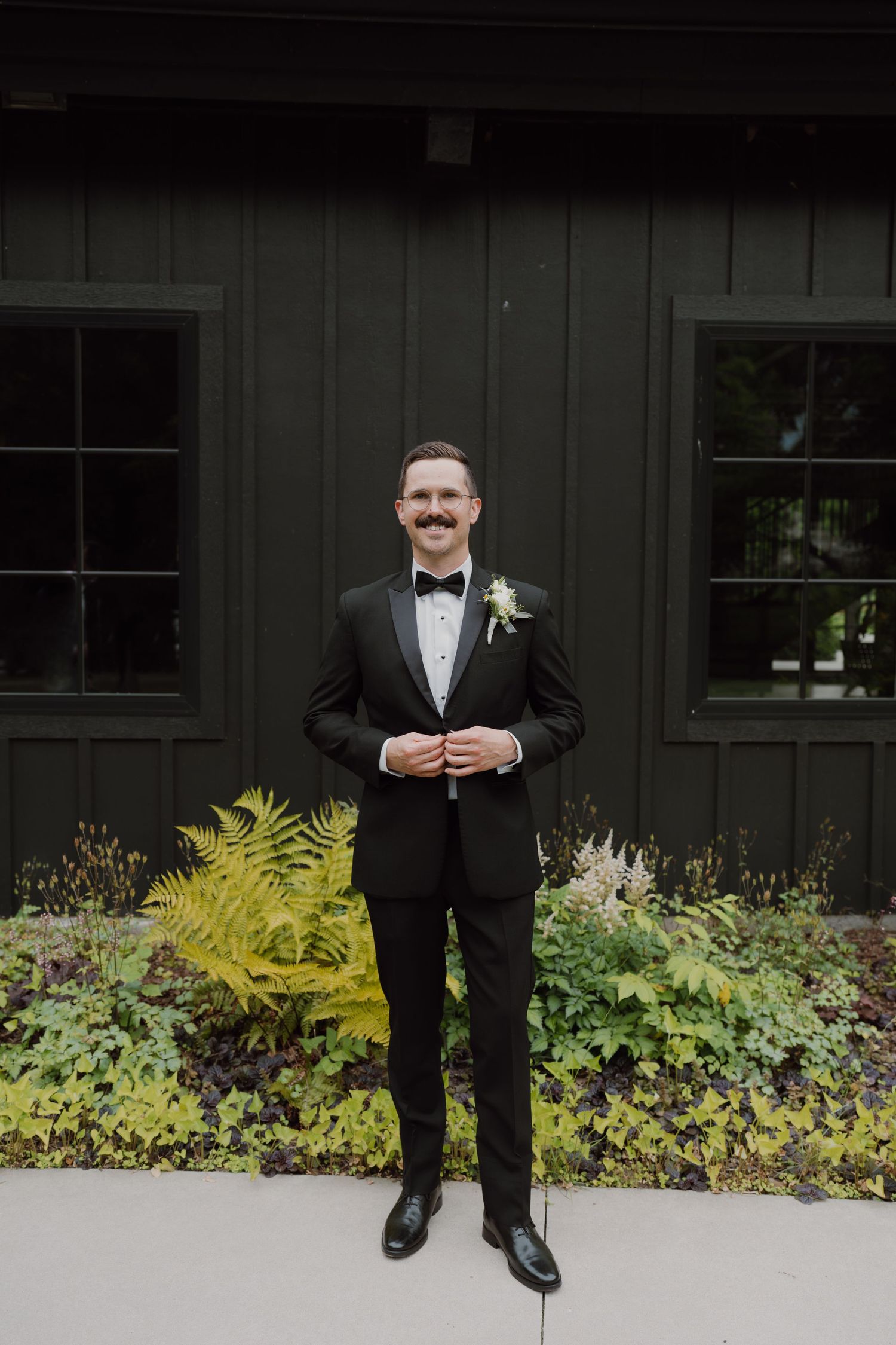 Groom in black tuxedo poses against dark wall with yellow ferns and garden foliage.