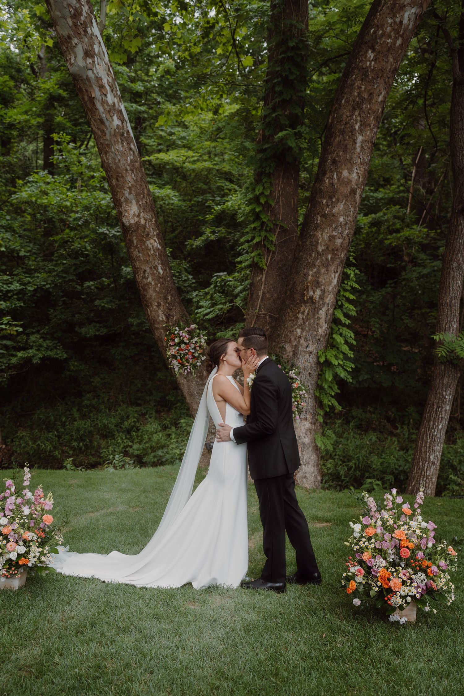 A romantic wedding scene unfolds between two trees adorned with vibrant flower arrangements in a forest setting.