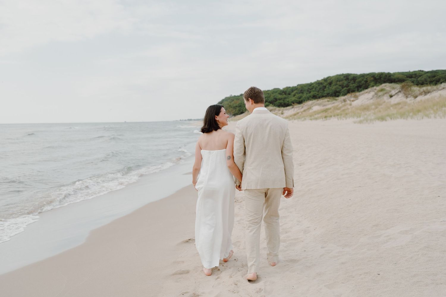 A couple takes a leisurely walk along a scenic beach with green vegetation in the background.