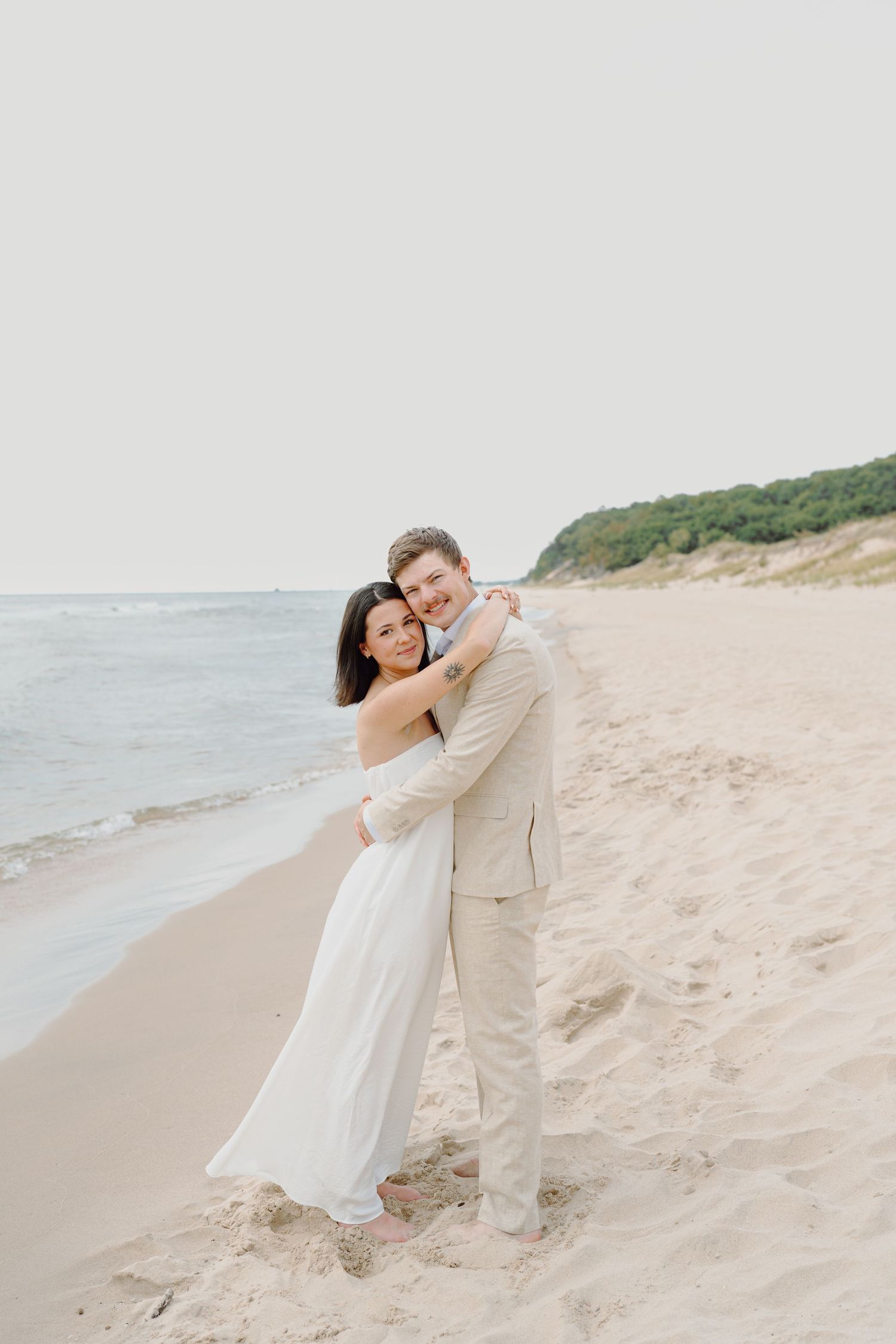 Couple in light colored clothing embrace on sandy beach with green cliffs in background.