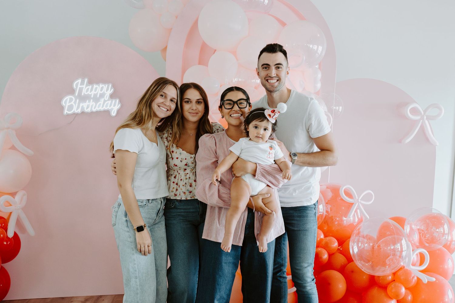 Group photo against pink and coral balloon display at birthday celebration.