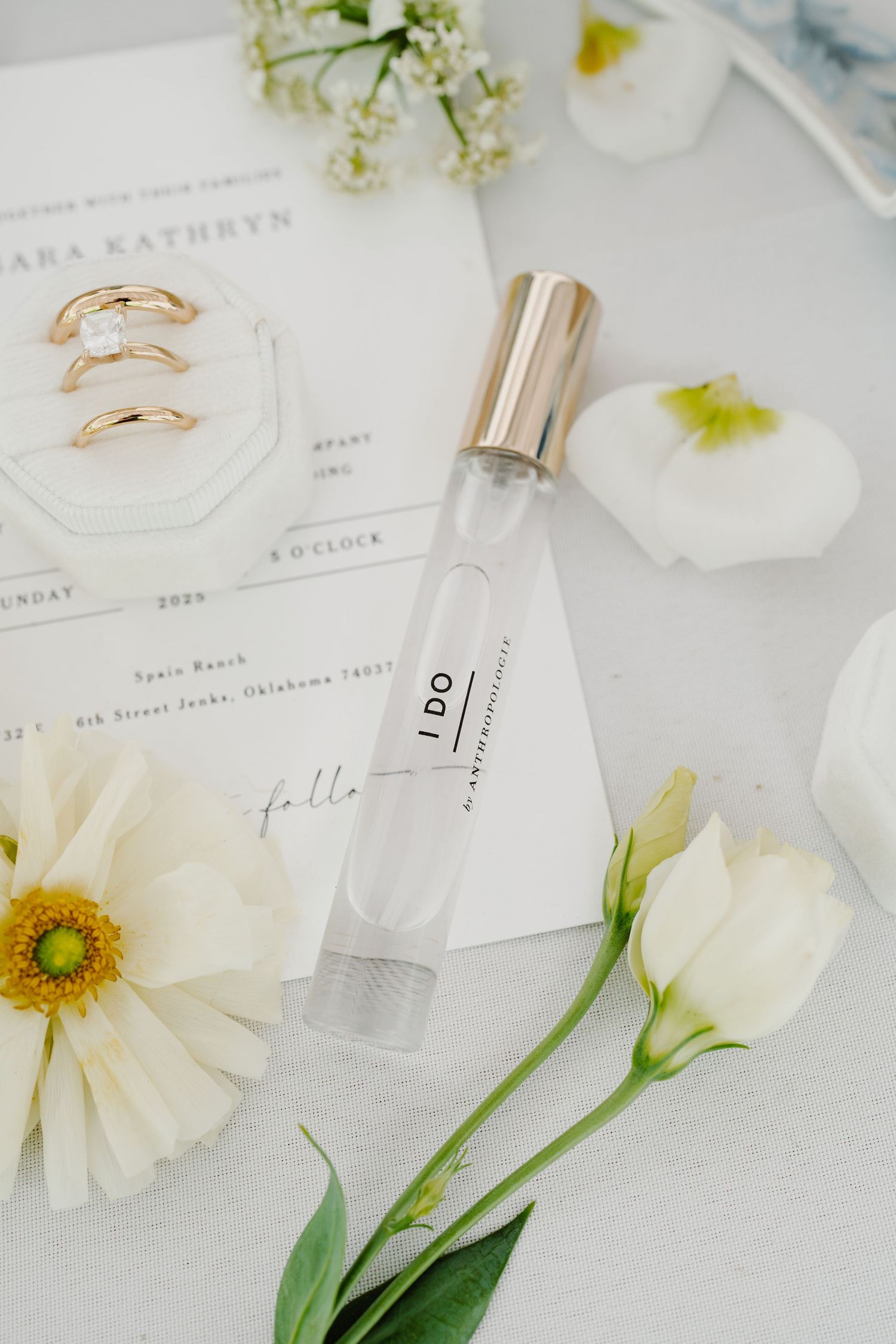 Wedding details including rings, perfume bottle, and white flowers arranged on a light surface.