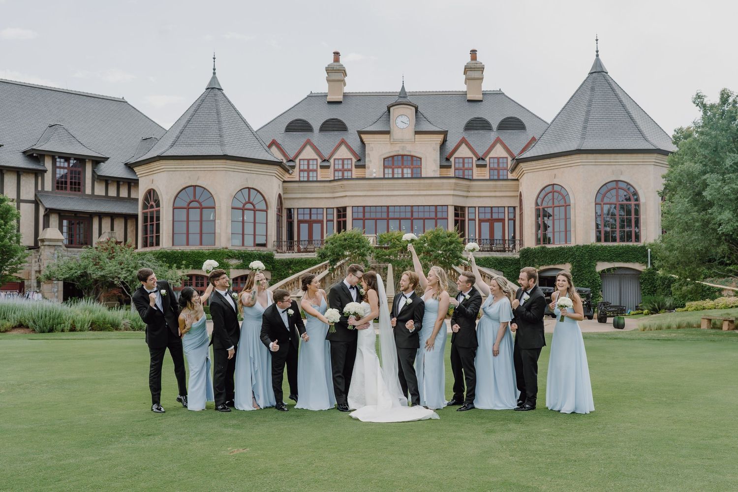 Wedding party members pose together on a lush green lawn outside an elegant stone mansion.