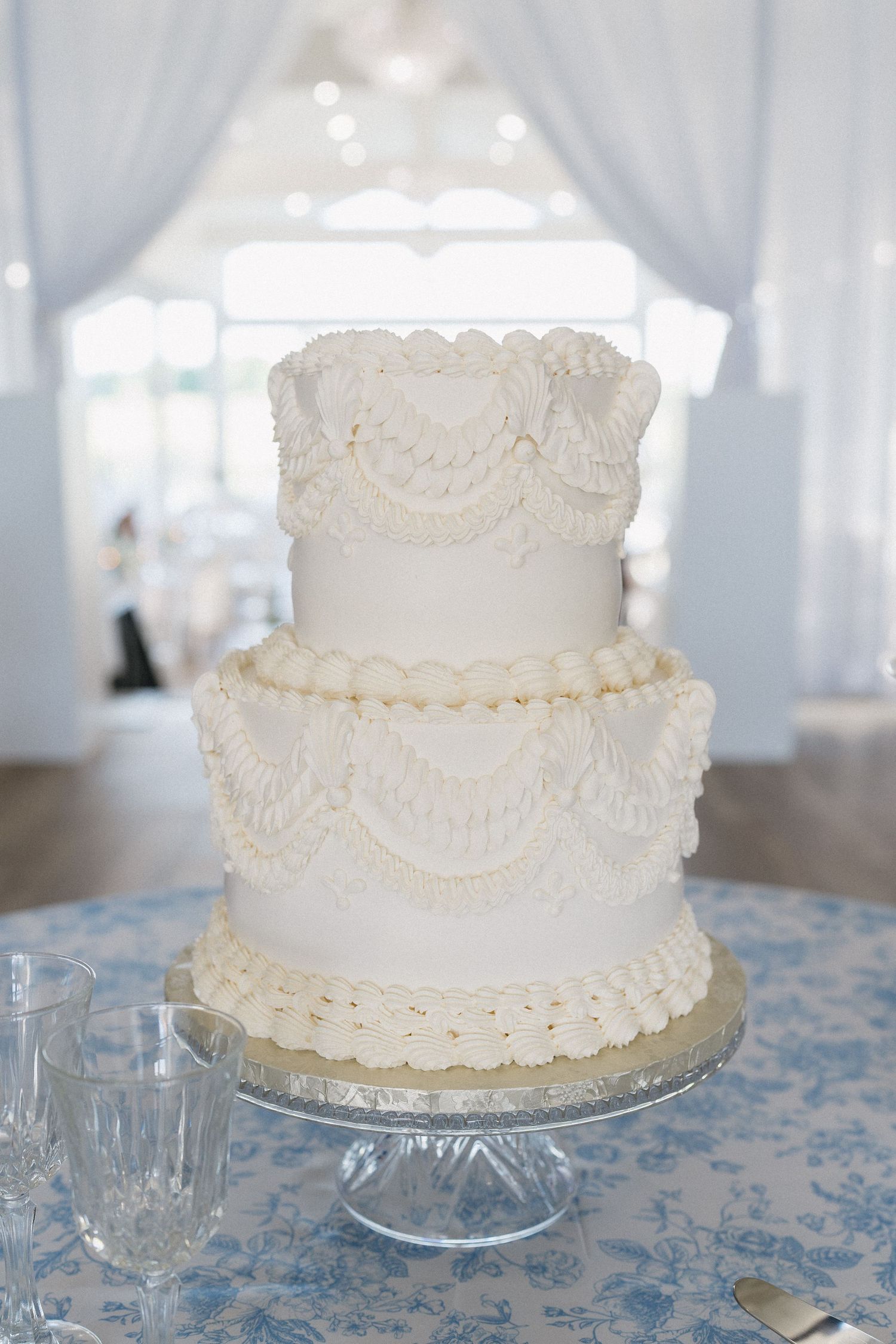 Elegant three-tier white wedding cake with intricate piped lace details on a glass stand with blue floral tablecloth.